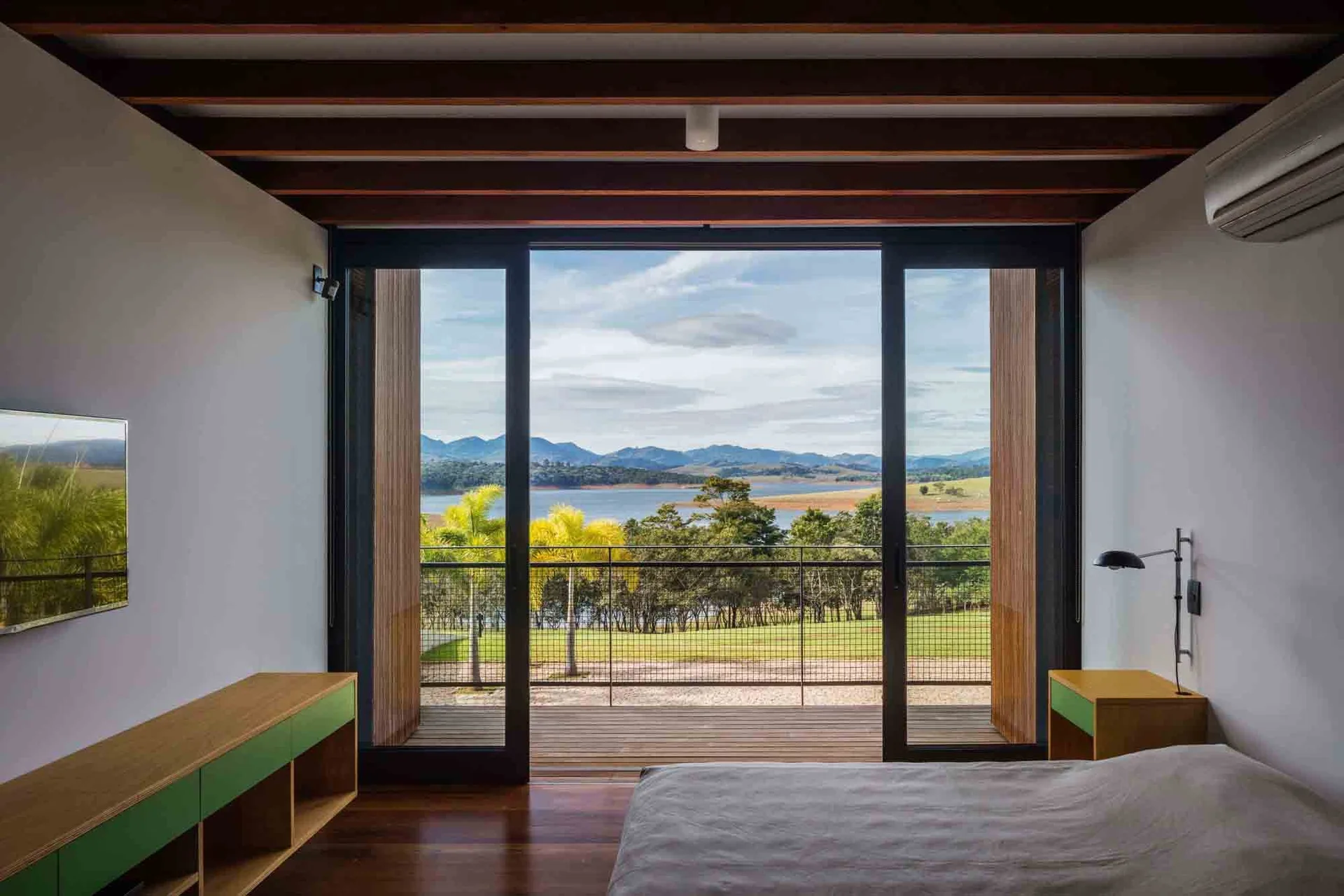 A minimalist bedroom features dark wood ceiling beams, a low wooden media console, and large sliding glass doors opening to a scenic lake view.