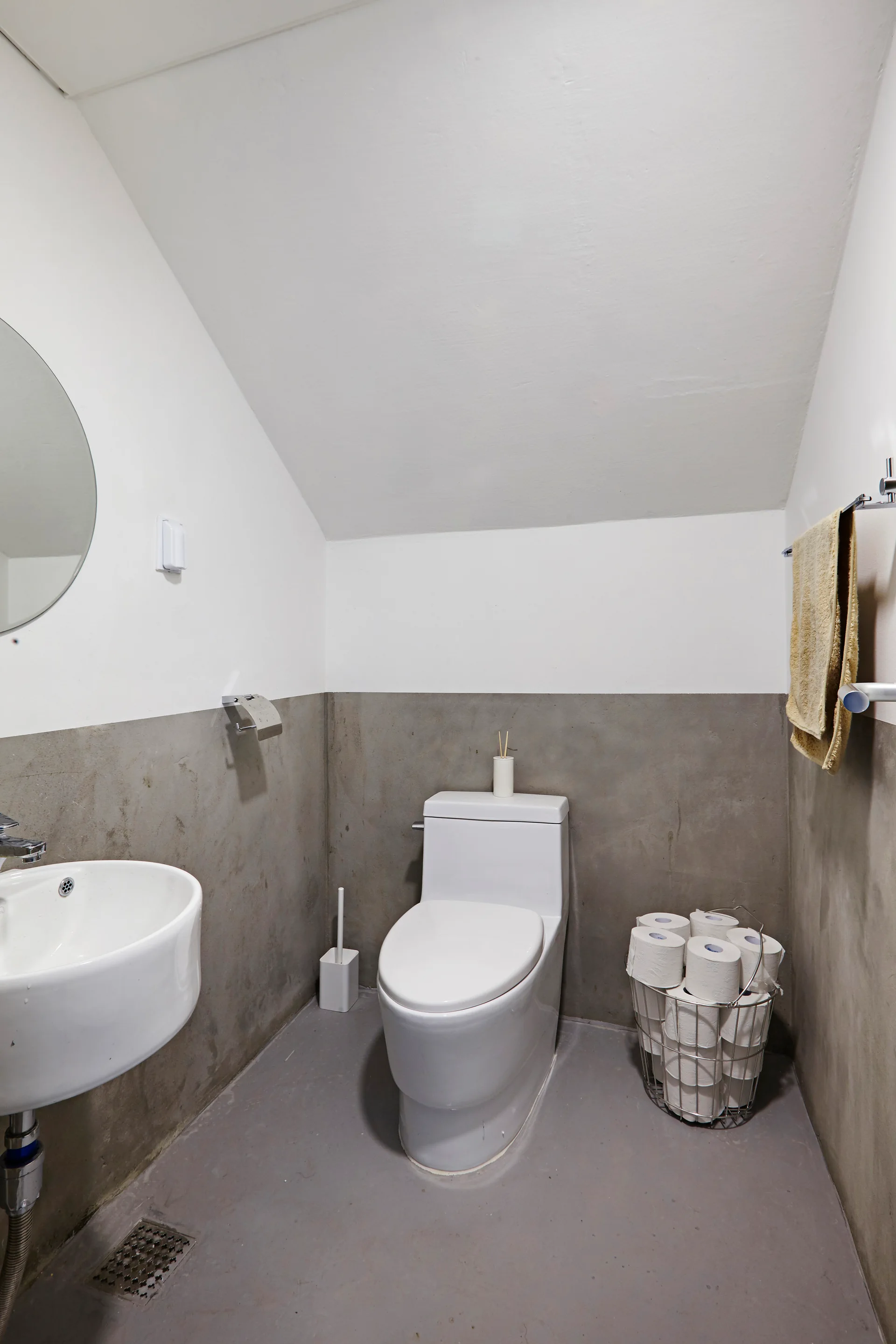 A compact powder room with a sloped ceiling features concrete-textured walls, a white toilet, a wall-mounted sink, and a wire storage basket.