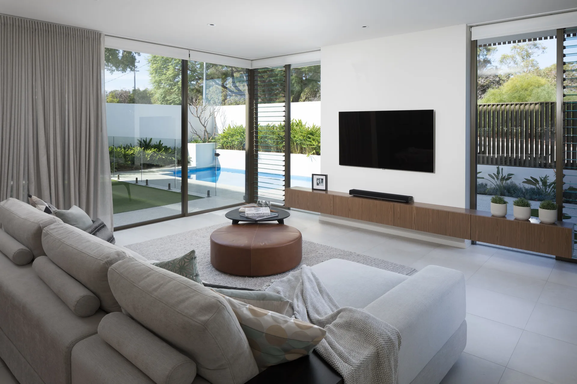 A modern living room features a grey sectional sofa, a round leather ottoman, and a floating wood console facing a swimming pool through large windows.