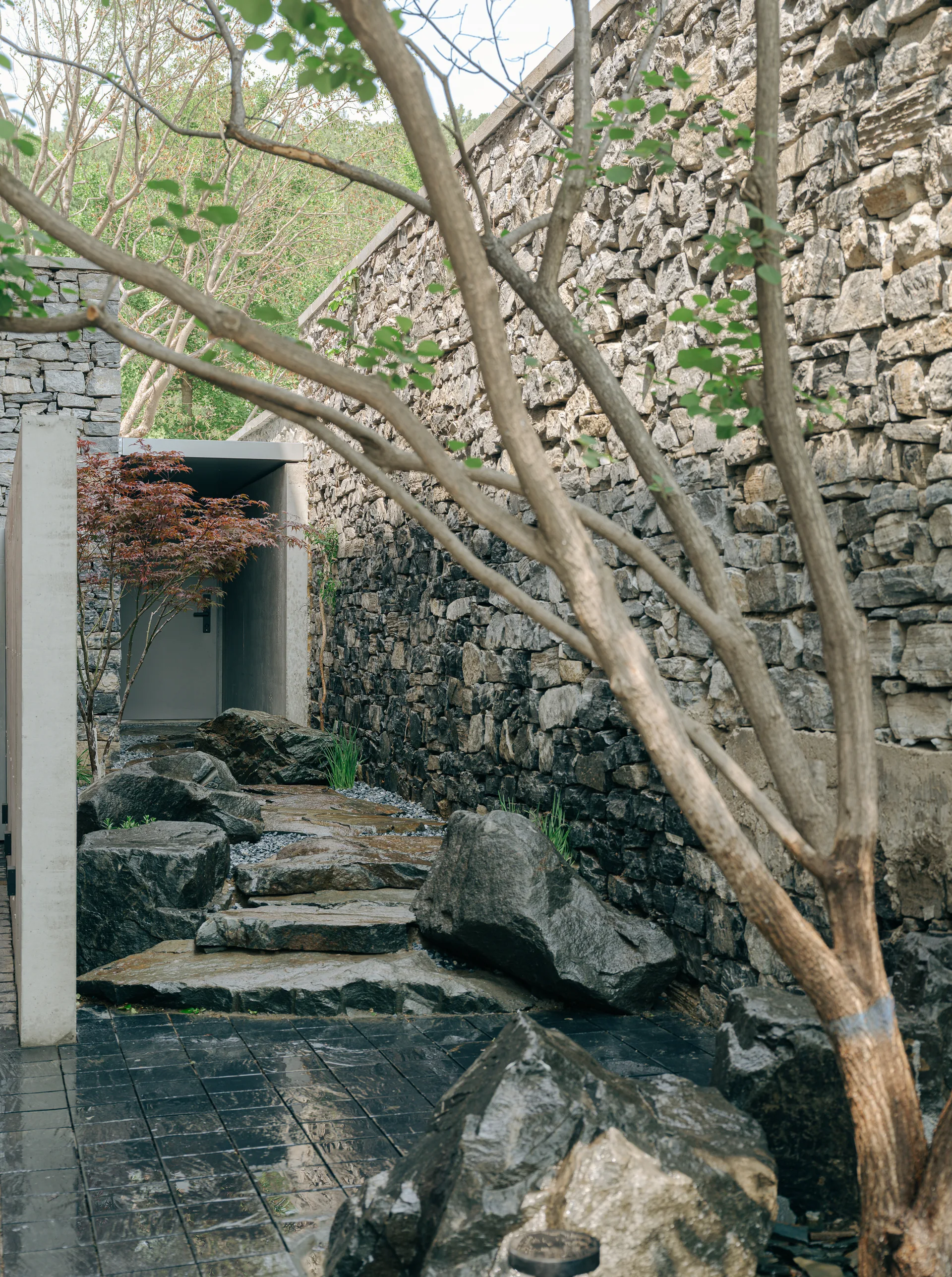 An outdoor garden path of large stepping stones winds alongside a tall, dry-stacked stone wall toward a concrete entryway.