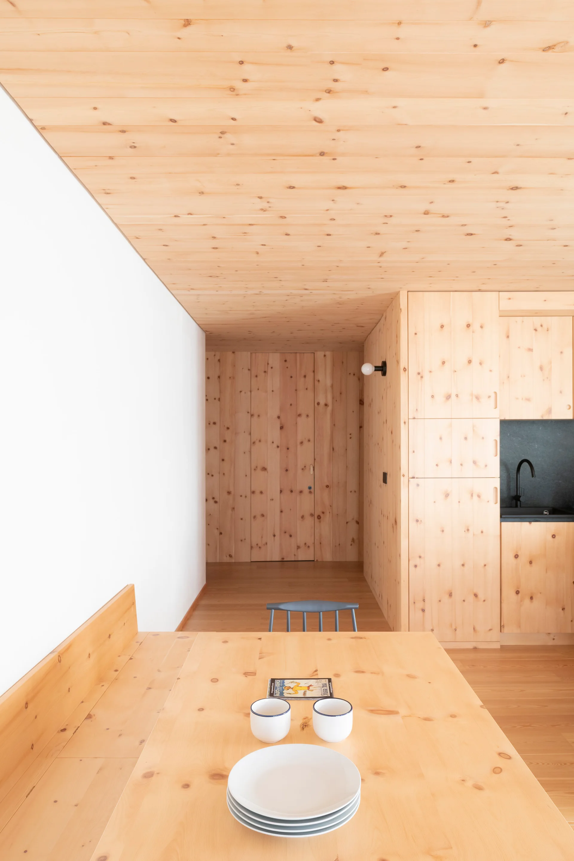 A wooden dining table sits before a minimalist kitchen with extensive knotty pine wall paneling, ceiling planks, and integrated cabinetry.