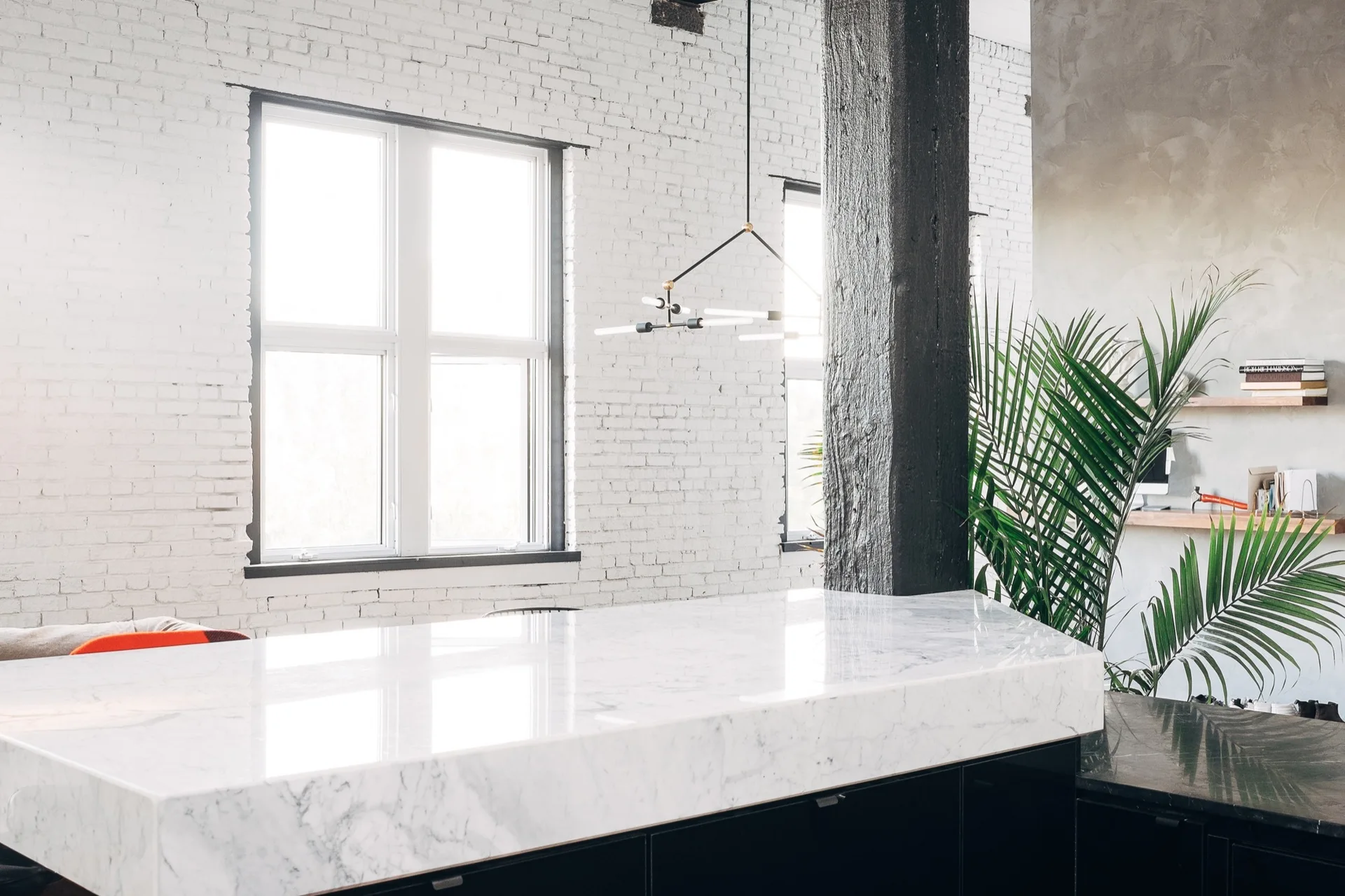 A modern loft kitchen features a polished white marble island, a dark structural pillar, and a large window set against a white painted brick wall.