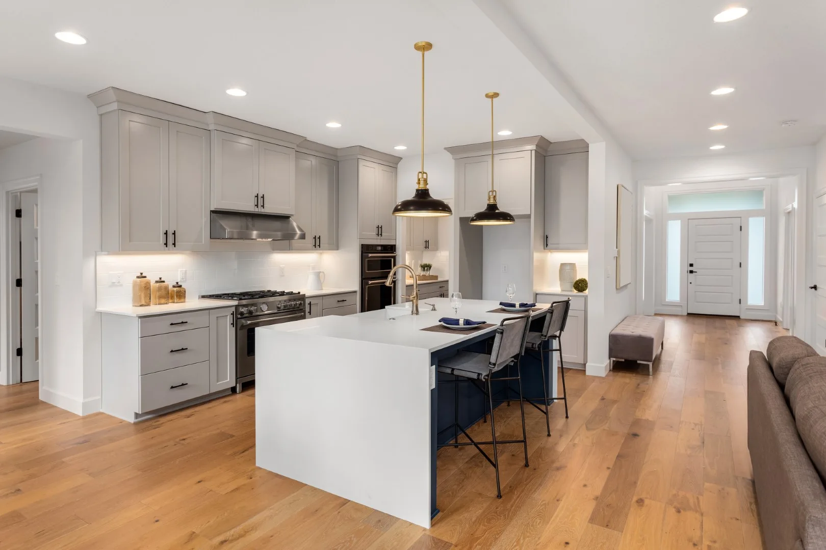 An open-concept kitchen features grey cabinets, a white and navy blue island with bar stools, and warm wood floors under dome pendant lights.