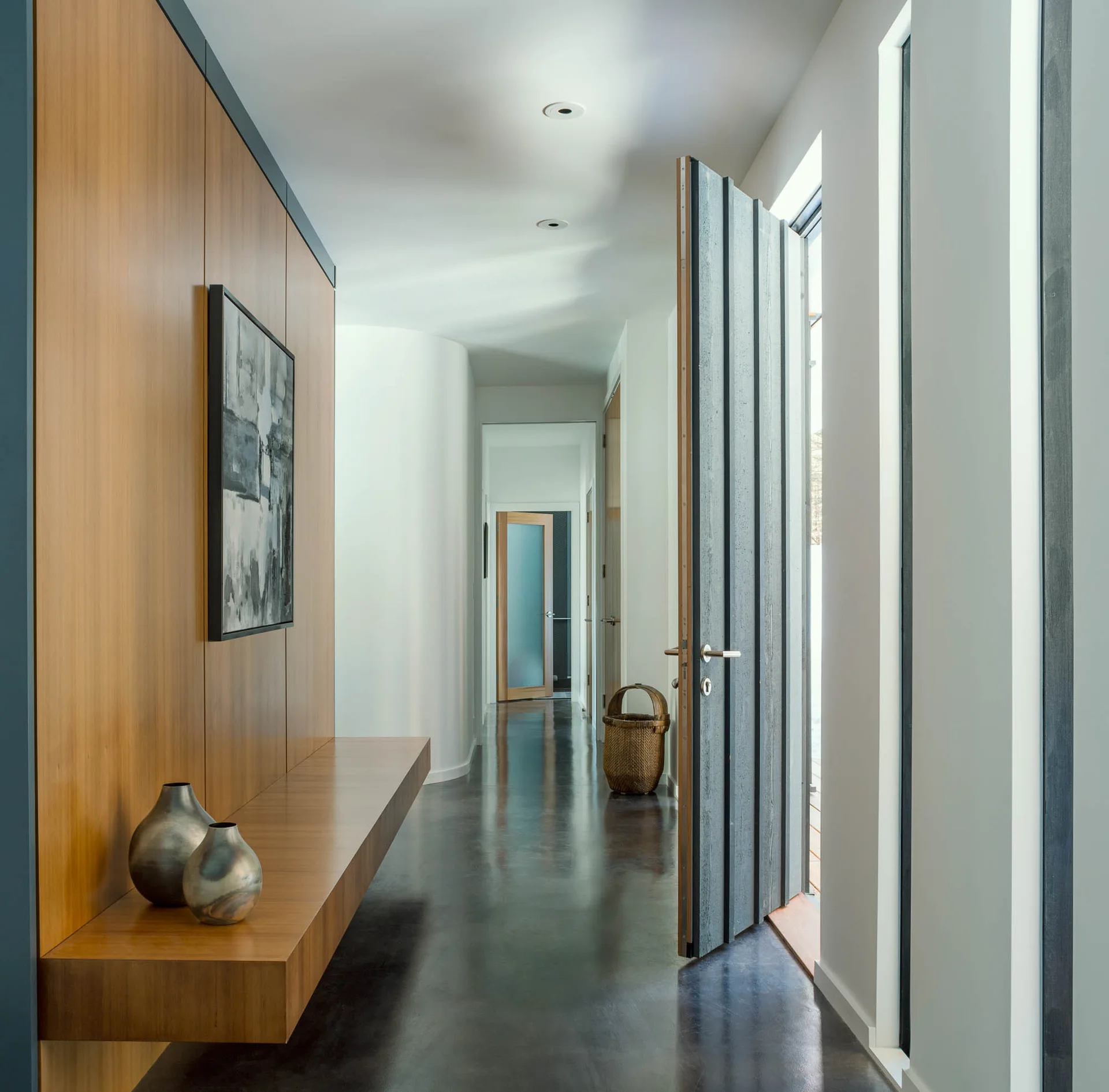 A modern hallway with a warm wood paneled wall, integrated floating wood bench, dark reflective glossy floor, and an open gray entry door.
