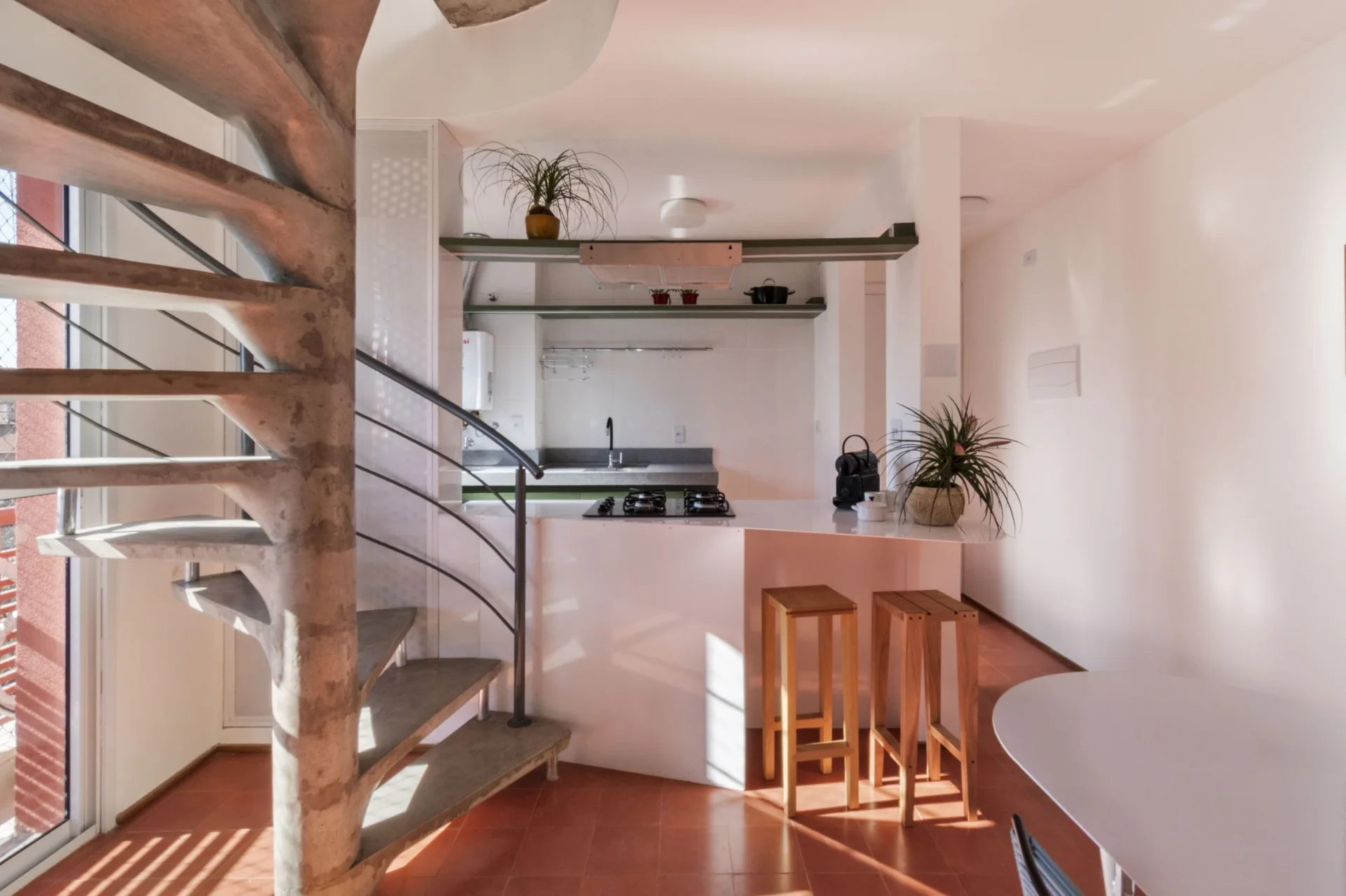 A concrete spiral staircase stands next to a white kitchen island with wooden bar stools on a terracotta tile floor.