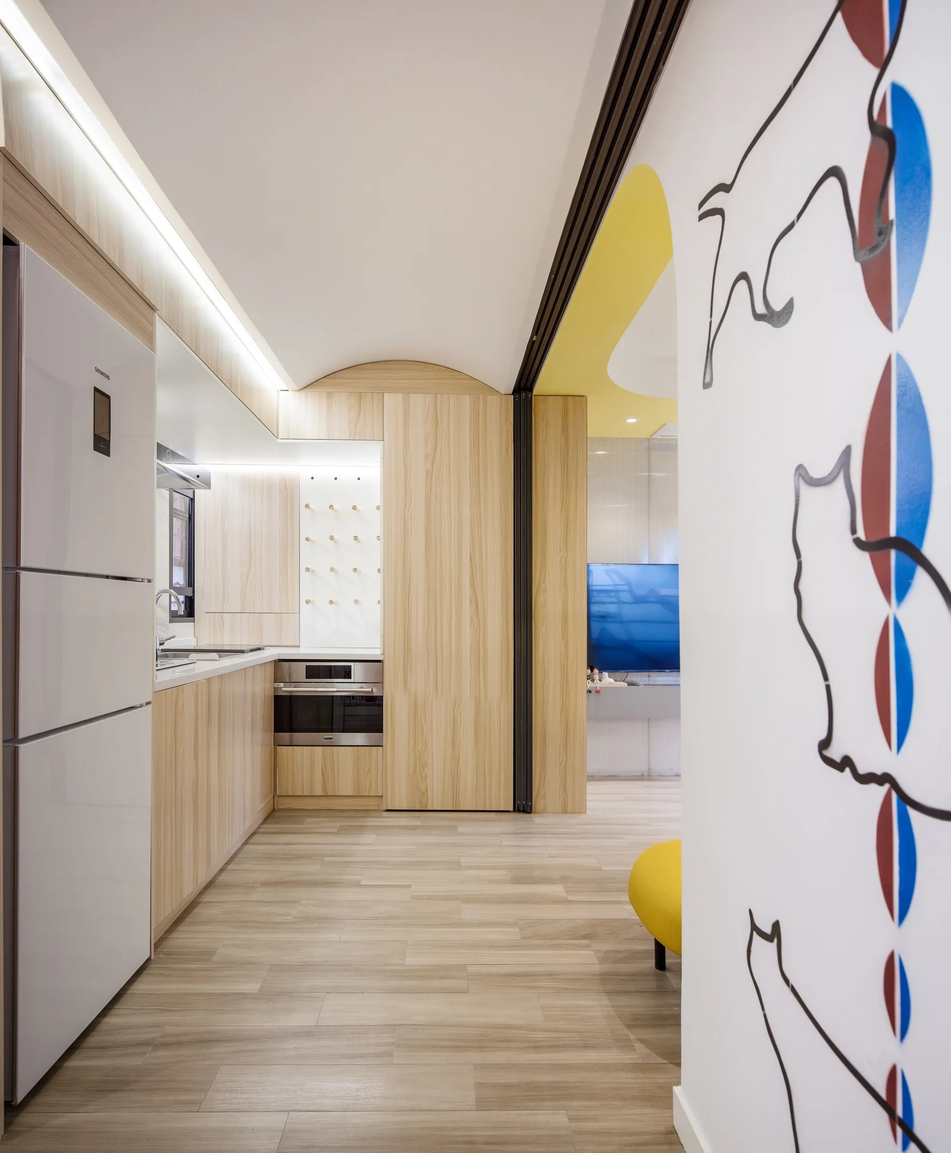 A modern kitchen features light wood cabinets, integrated LED lighting, and a bright yellow arched doorway leading to an adjacent room.