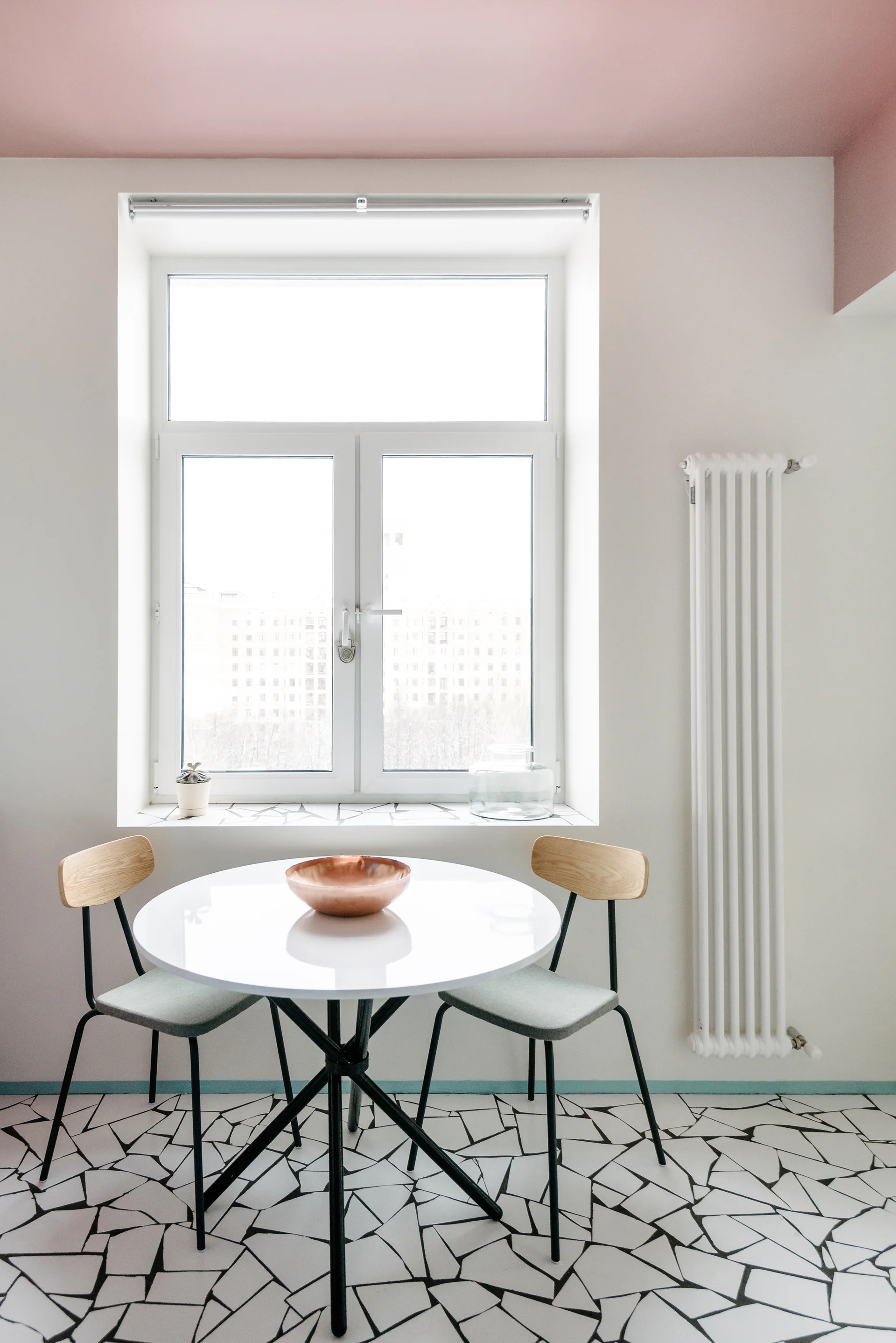 A minimalist dining area features a white round table, two wood-backed chairs, a black-and-white mosaic floor, and a pastel pink painted ceiling.