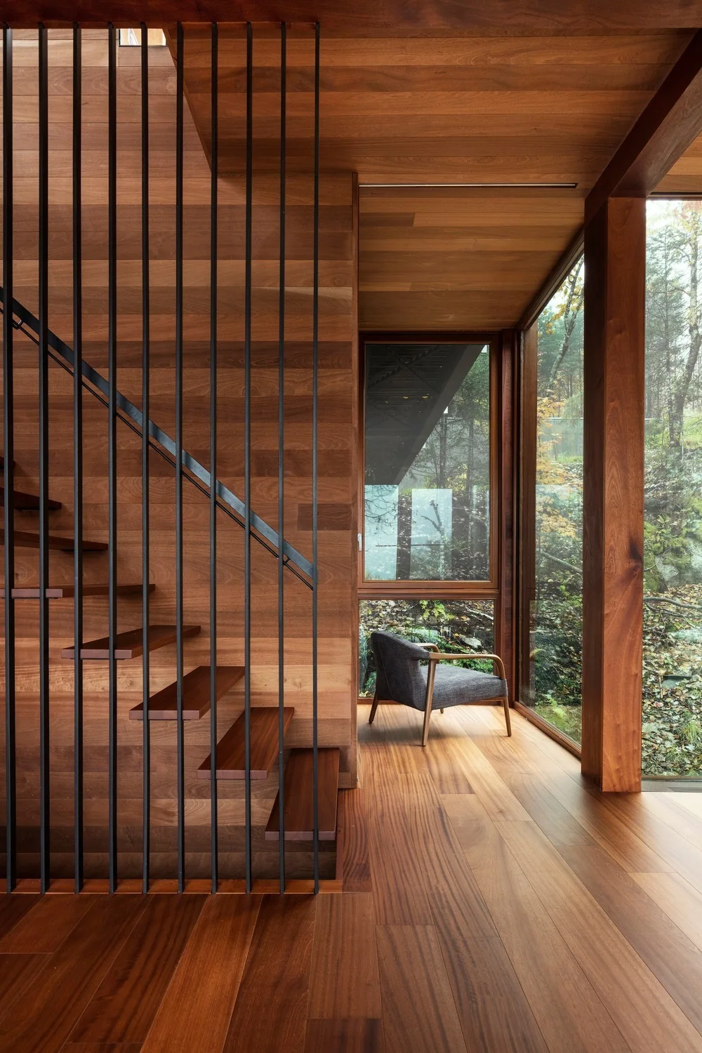 A wooden staircase with open treads and vertical metal spindles sits next to wood-paneled walls and a large window looking out to a forest.