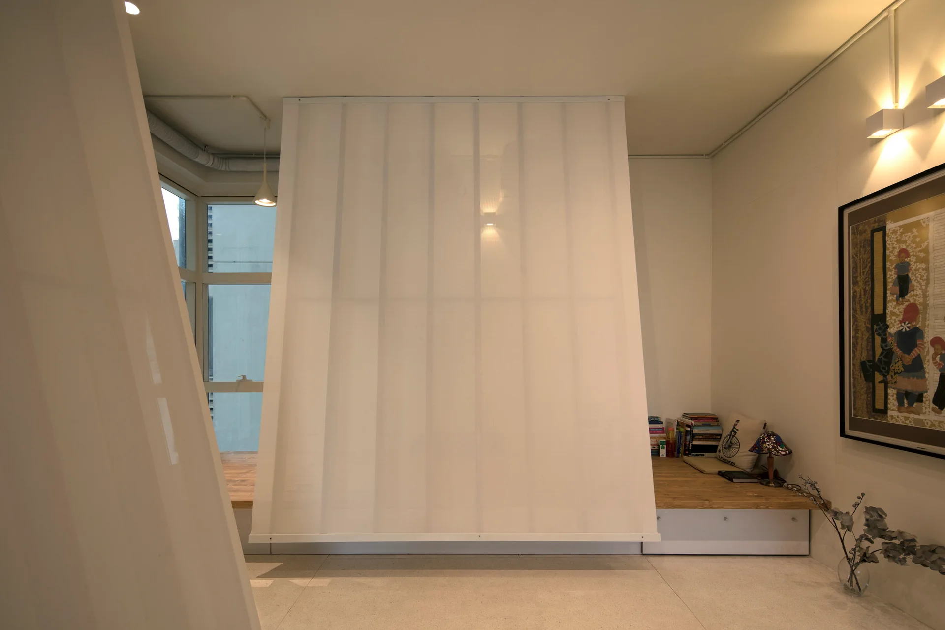 A white hanging fabric panel partition divides a minimalist room featuring a raised wooden platform bench near a window and framed artwork on the wall.
