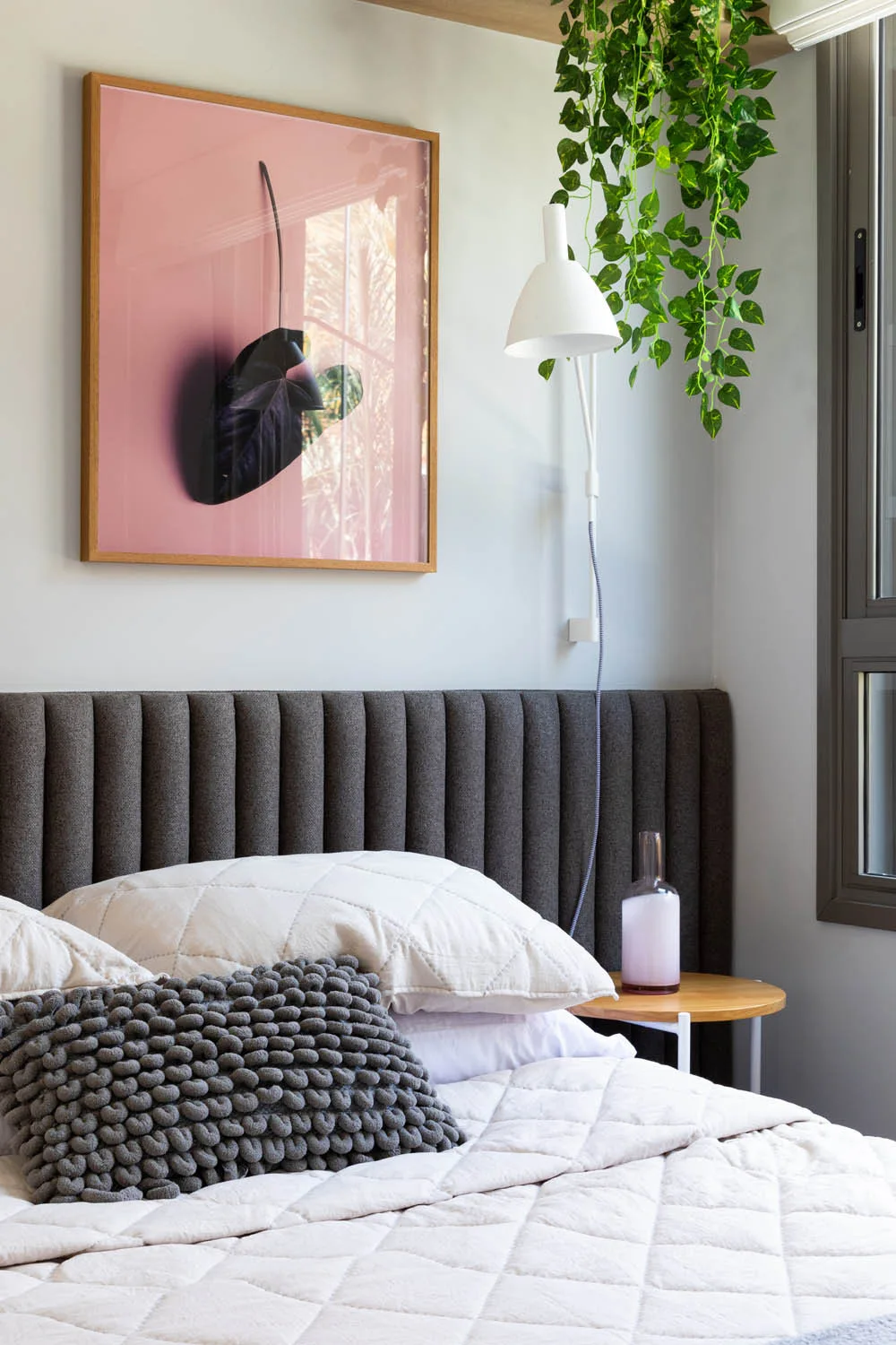 A cozy bedroom corner featuring a charcoal gray channel-tufted headboard, quilted white bedding, a white wall-mounted lamp, and a framed pink art print.