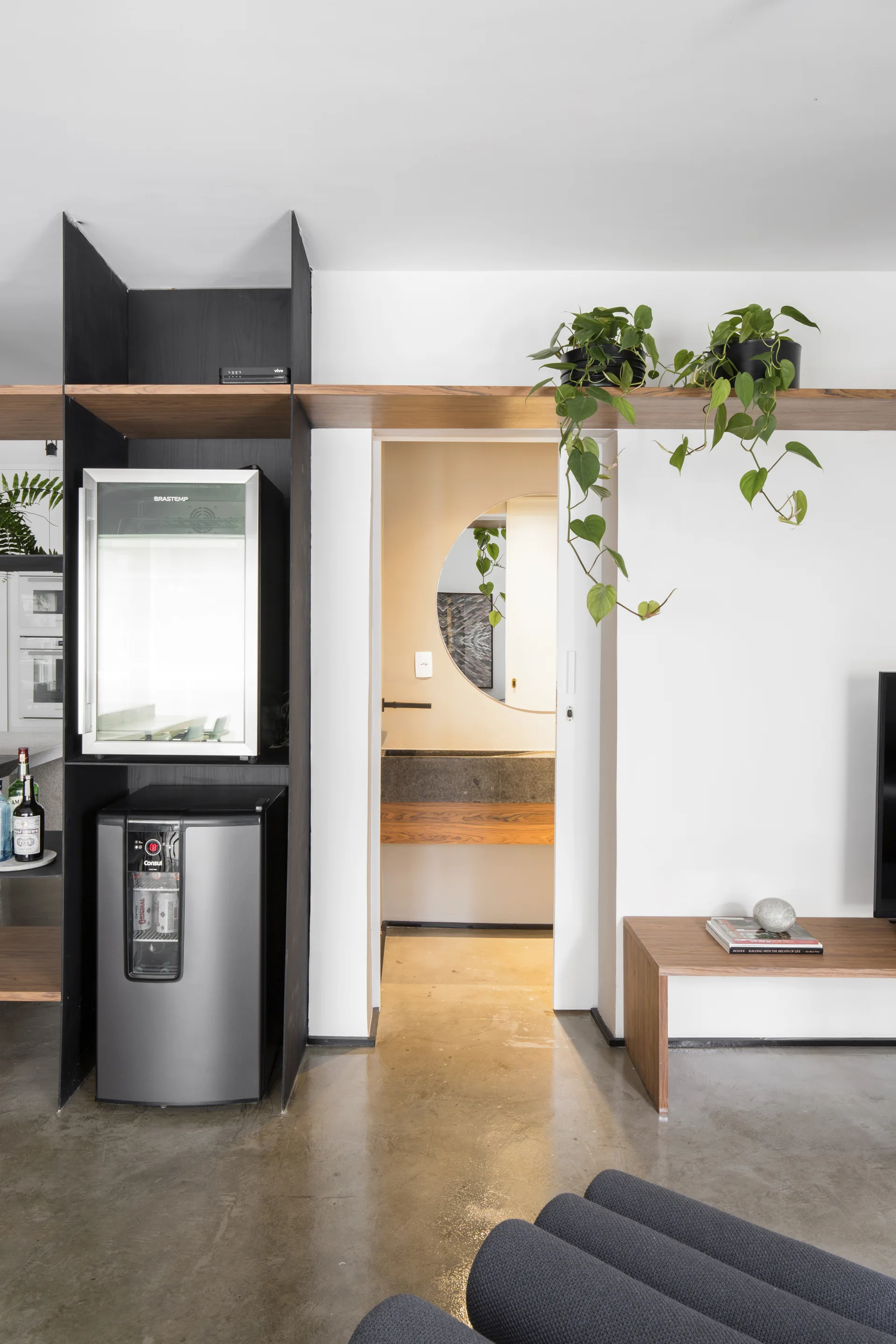 A modern interior features a dark open shelving unit with a beverage cooler next to a doorway, a wooden floating shelf, and polished concrete floors.