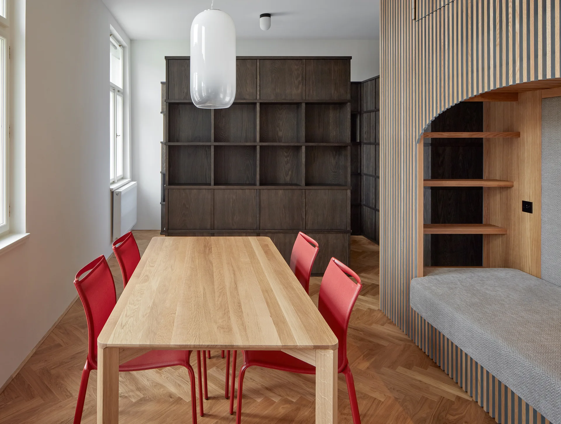 A light wood dining table with four red chairs stands near a dark wood bookcase and a built-in wall niche clad in vertical wood slats.