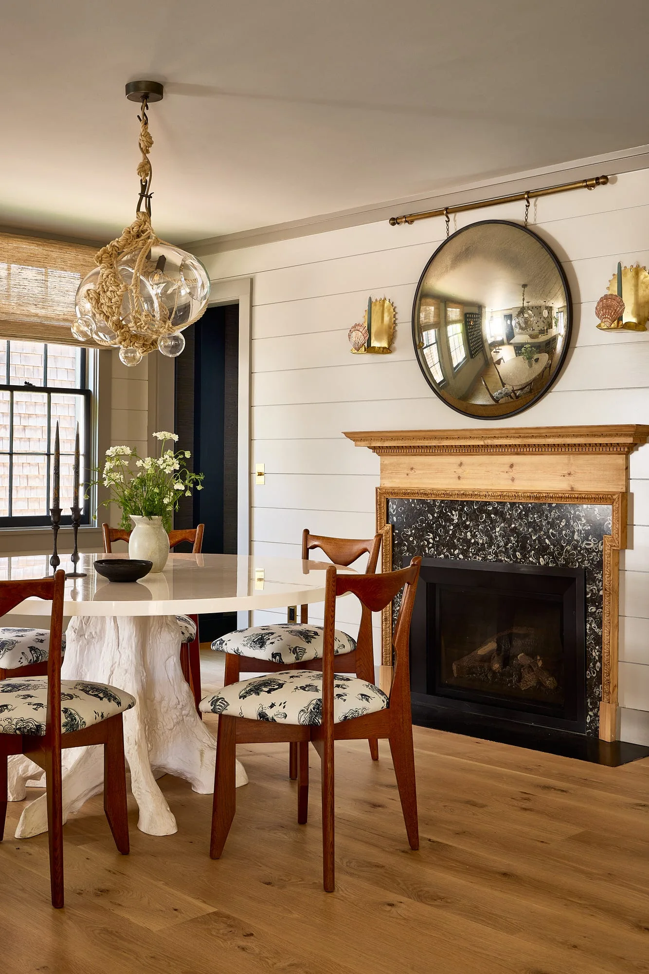 A bright dining room features a white sculptural table, patterned wooden chairs, a rope pendant light, and a fireplace with a wood mantel.