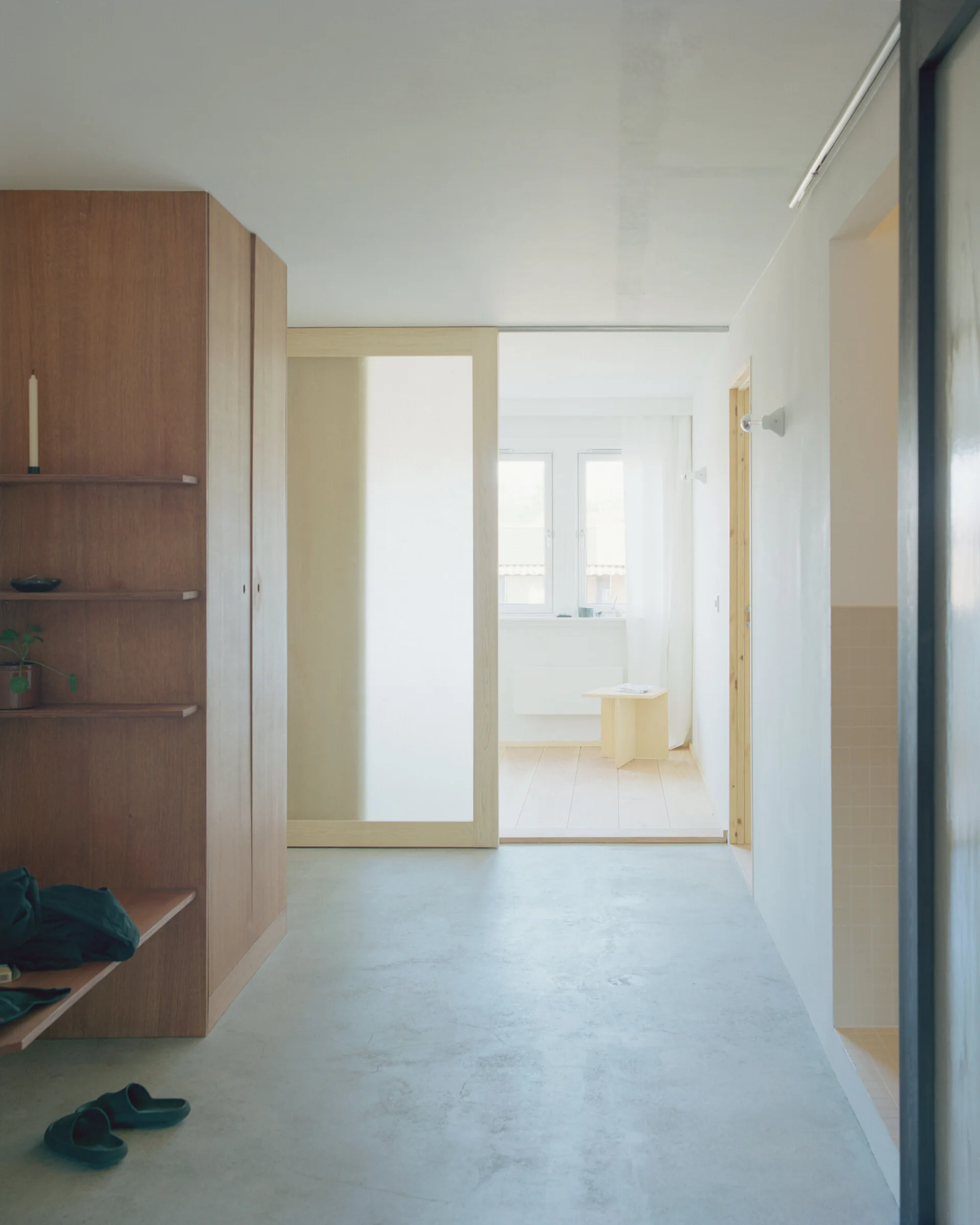 A minimalist hallway features a wood storage cabinet, gray concrete floor, and a wooden sliding door with a frosted glass panel leading to a sunlit room.