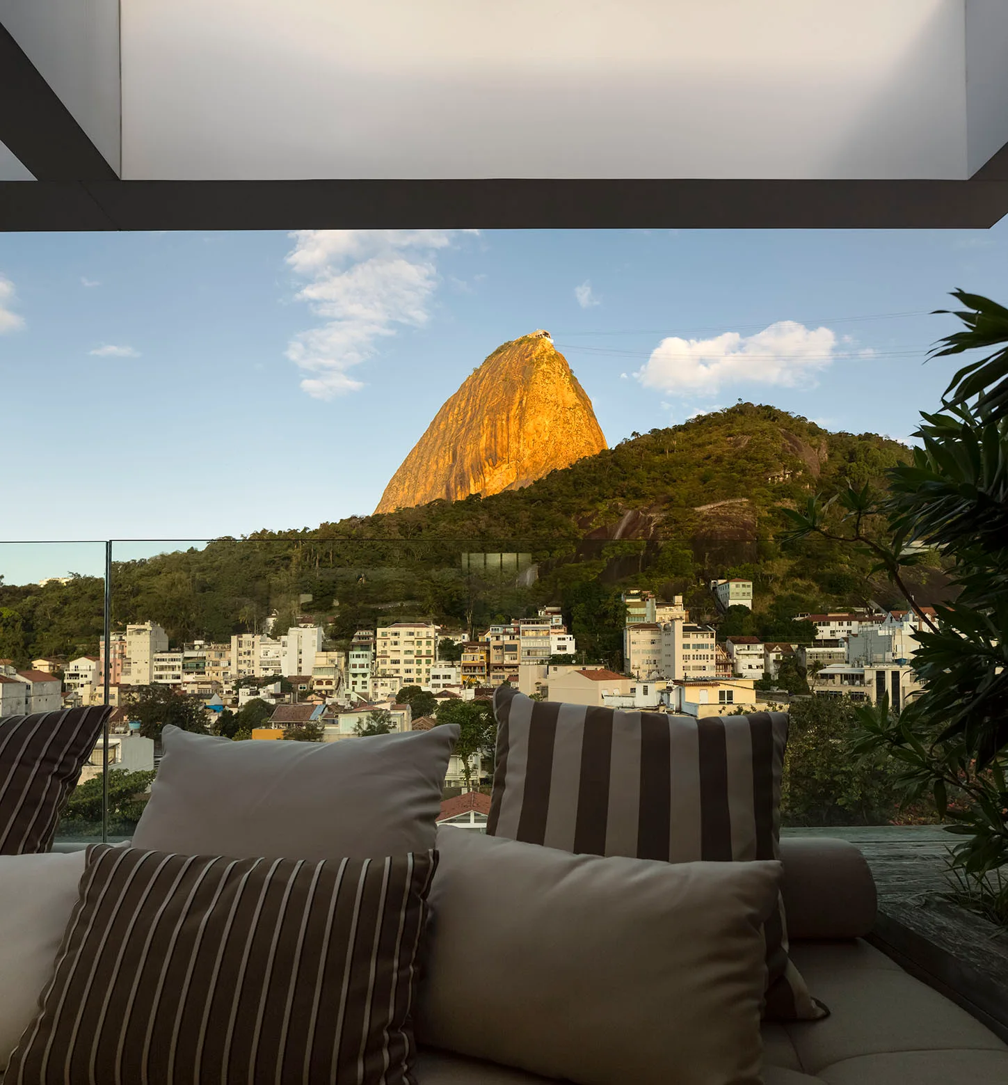 An outdoor terrace features a neutral cushioned sofa with striped pillows behind a glass railing overlooking a mountain landscape.
