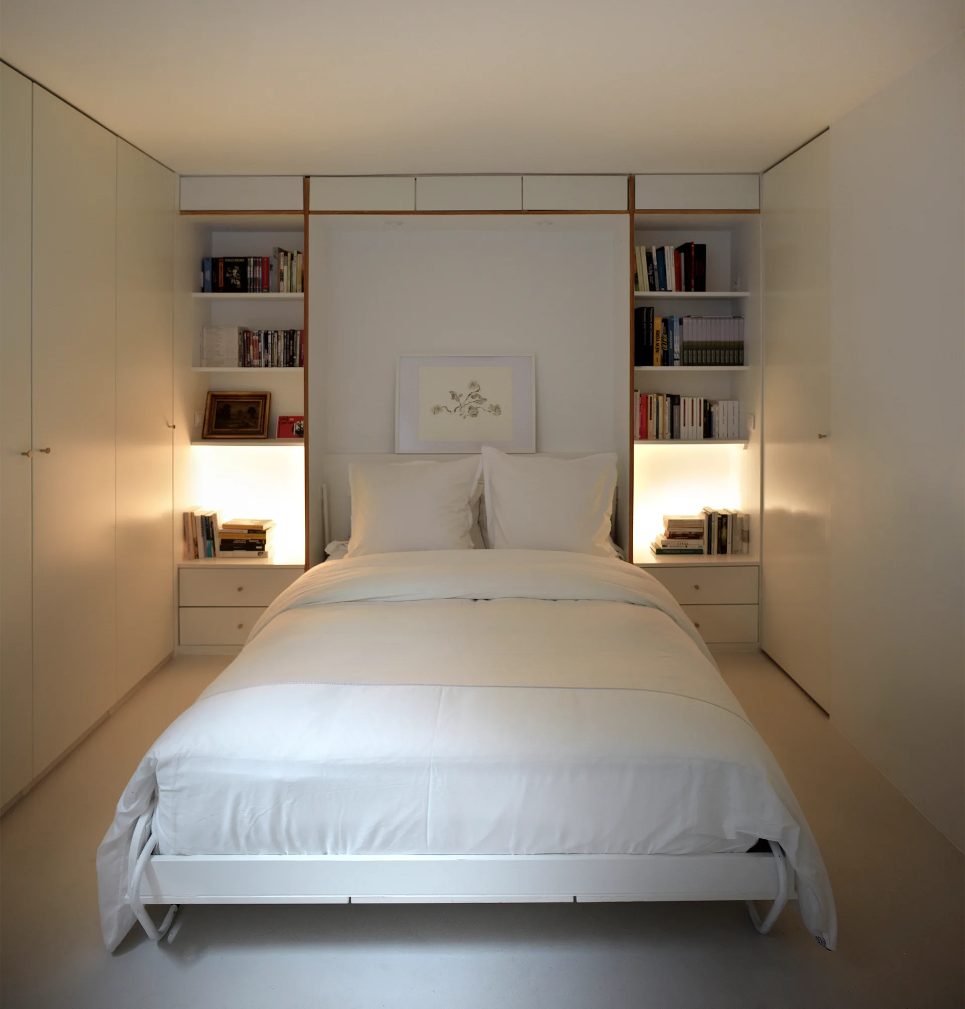 A minimalist white bedroom features a centered bed flanked by symmetrical built-in wardrobes, integrated shelves, and warm glowing undershelf lighting.