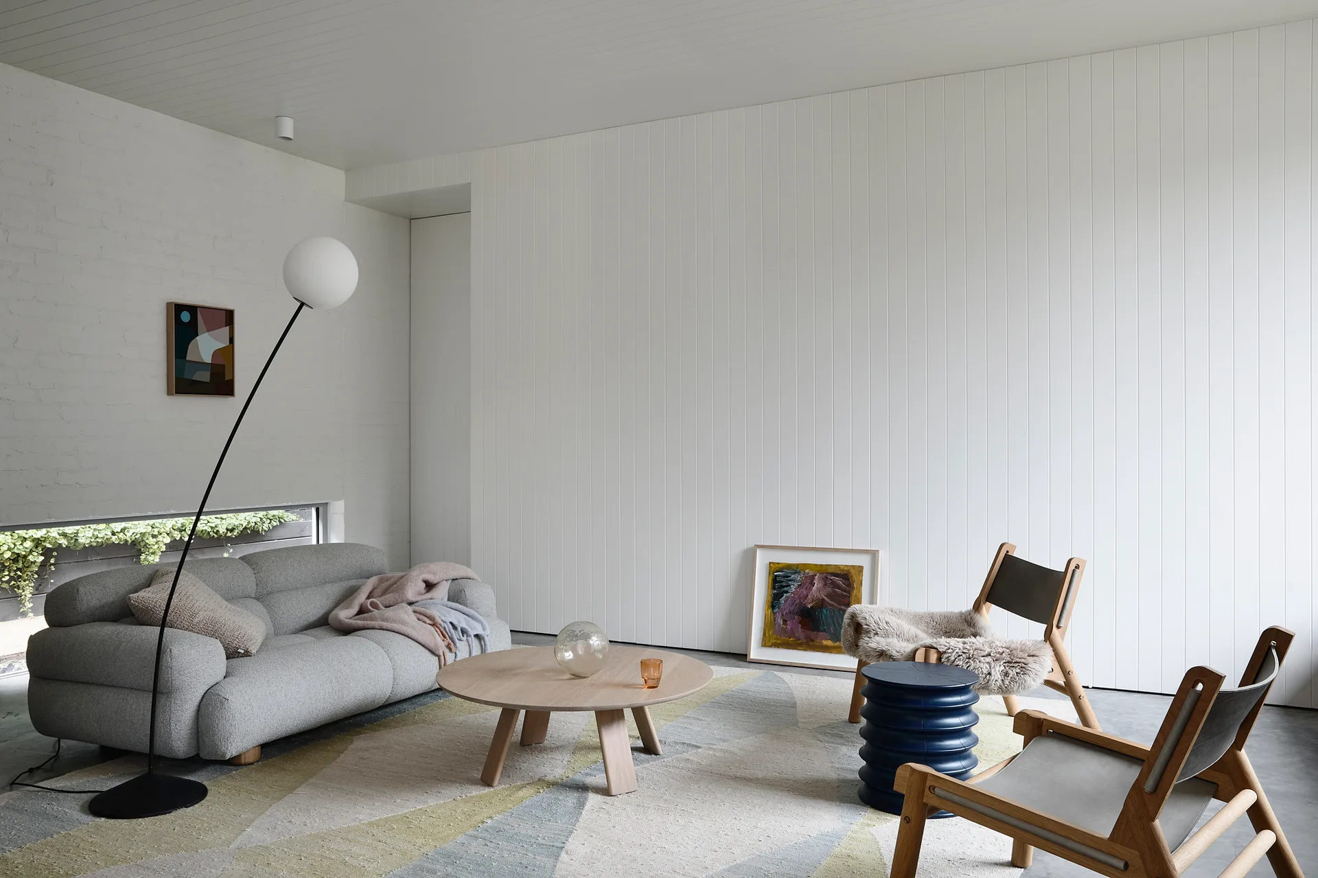 A modern living room with a grey sofa, wooden armchairs, and a round coffee table on a geometric rug against vertical white paneling.