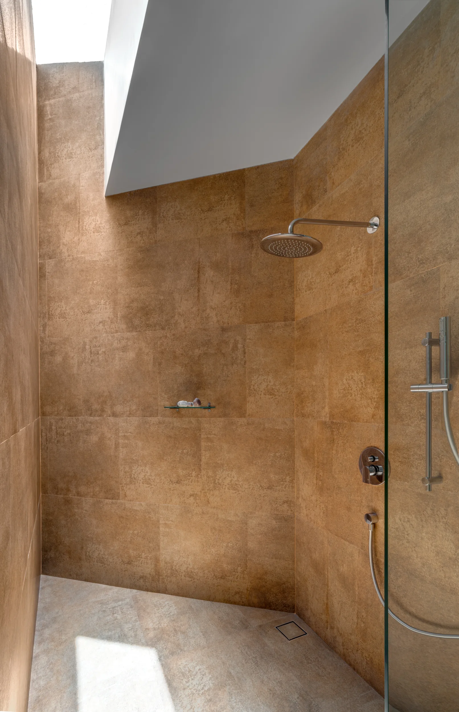 A minimalist walk-in shower features warm brown textured wall tiles, a glass partition, and bright natural light coming from an overhead skylight.