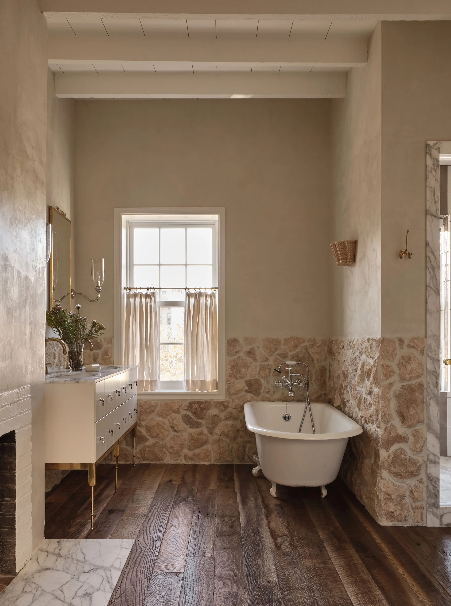 A rustic bathroom features a white clawfoot bathtub, a modern vanity with gold legs, stone wainscoting, and dark wood plank floors.