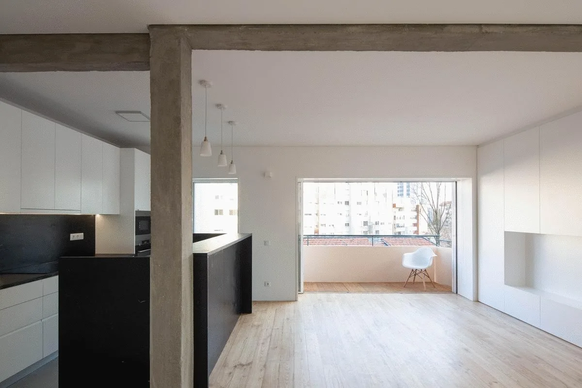 An open-plan living area and minimalist kitchen feature white walls, a black island, and an exposed raw concrete ceiling beam.