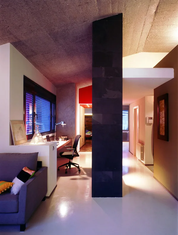 A modern home office with a built-in desk and chair is separated from a sofa area by a prominent black tiled column under an exposed concrete ceiling.