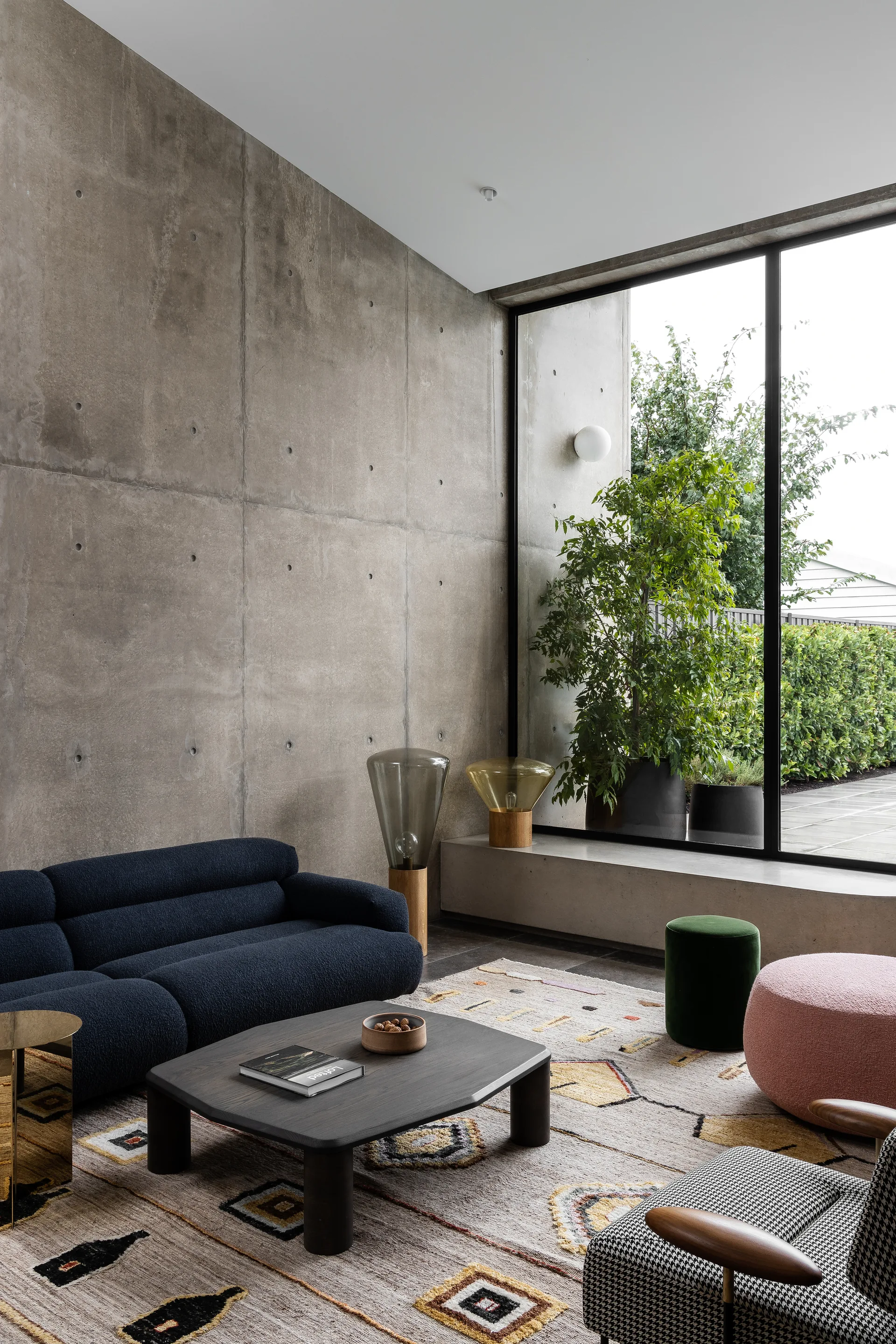 A modern living room features an exposed concrete wall, a navy blue sofa, a dark wood coffee table, and a large floor-to-ceiling window.