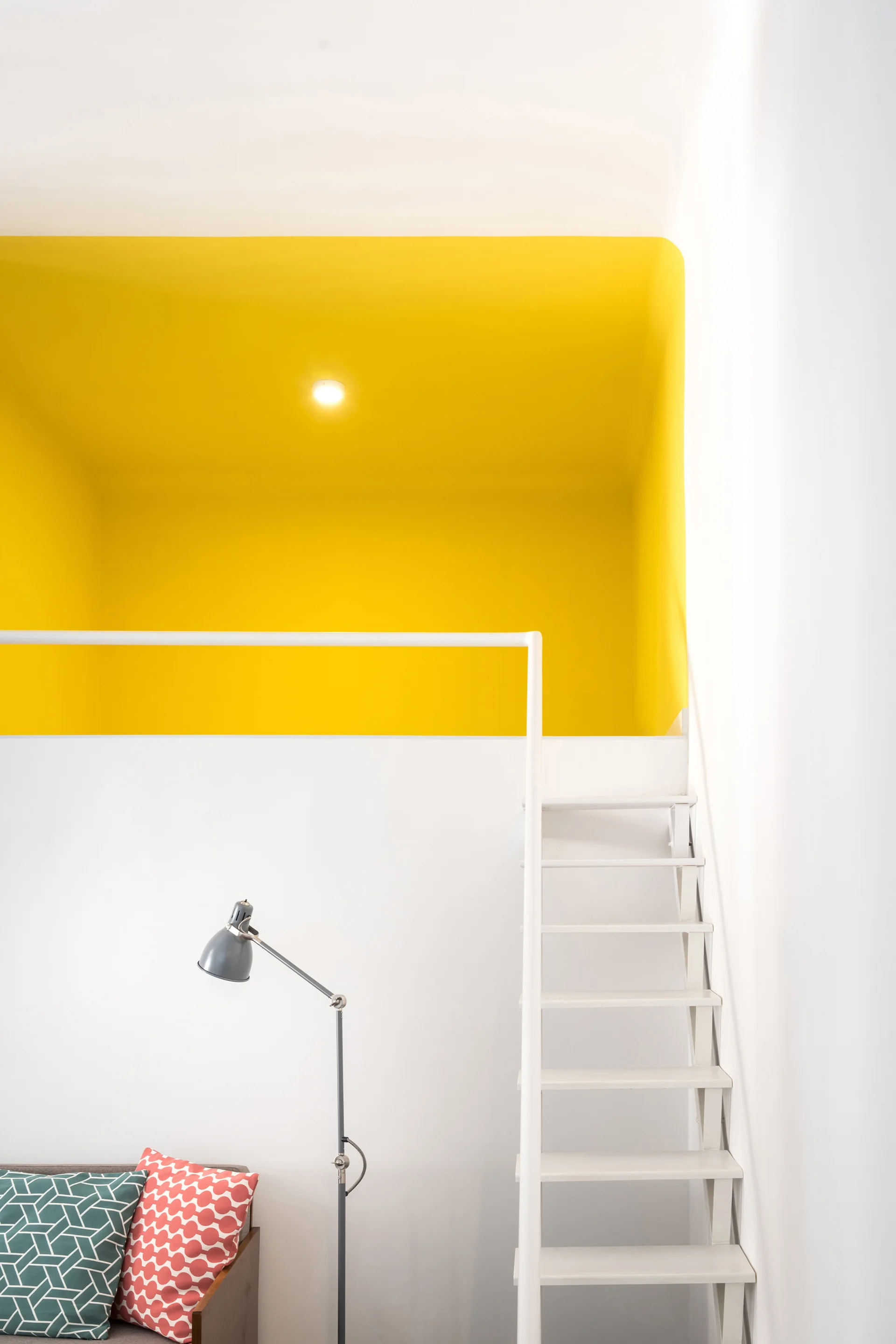 A white metal ladder staircase leads up to a mezzanine loft painted in solid bright yellow, set above a couch with geometric patterned pillows.