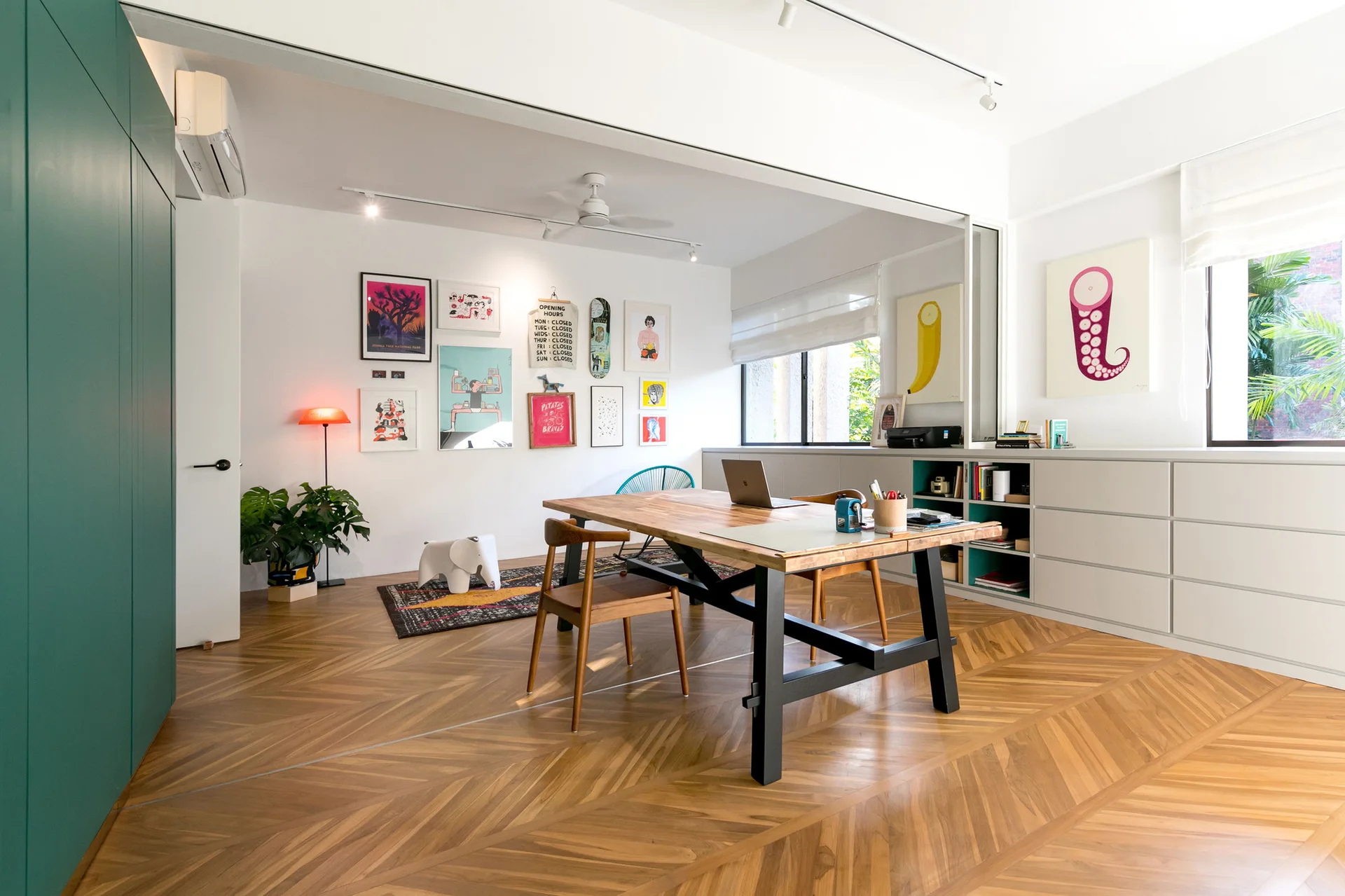 A bright home office features a wooden work table, herringbone wood floors, a colorful gallery wall, and a low white built-in storage unit.