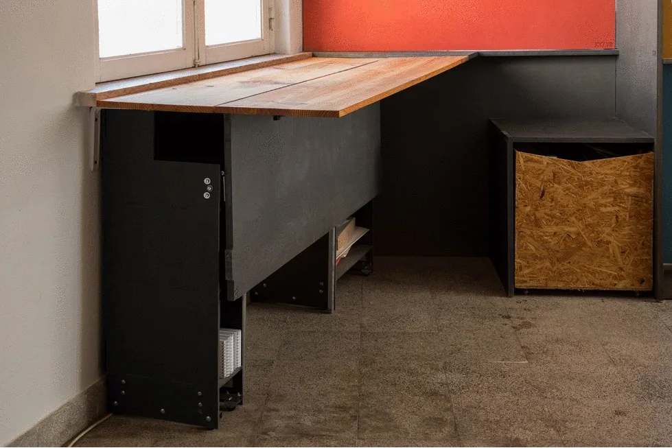 A wall-mounted wooden desk stands beneath a window, paired with a black metal base and a textured OSB storage bin against a red accent wall.