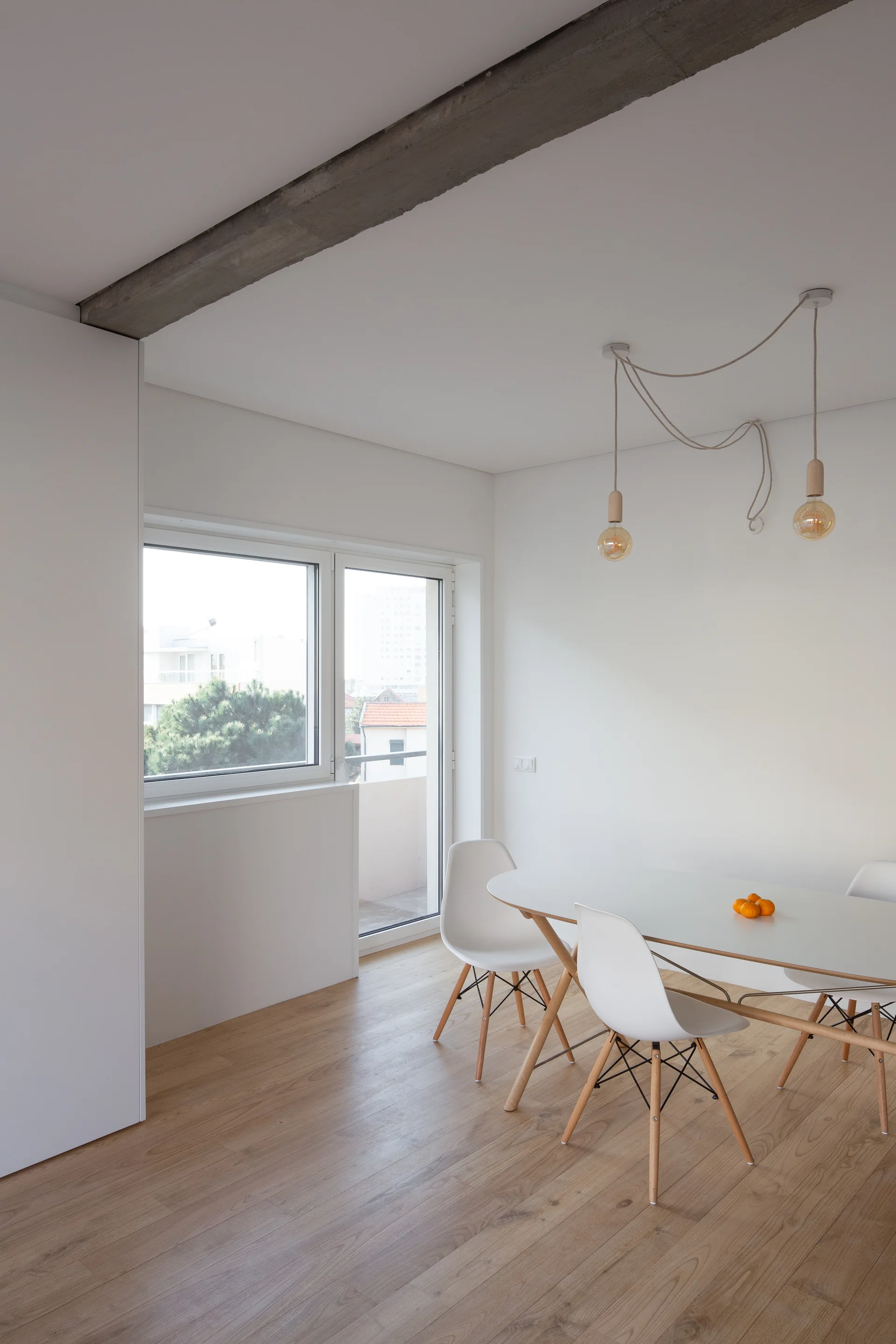 A minimalist dining room features a white oval table and chairs on wood flooring beneath a swagged pendant light and concrete ceiling beam.