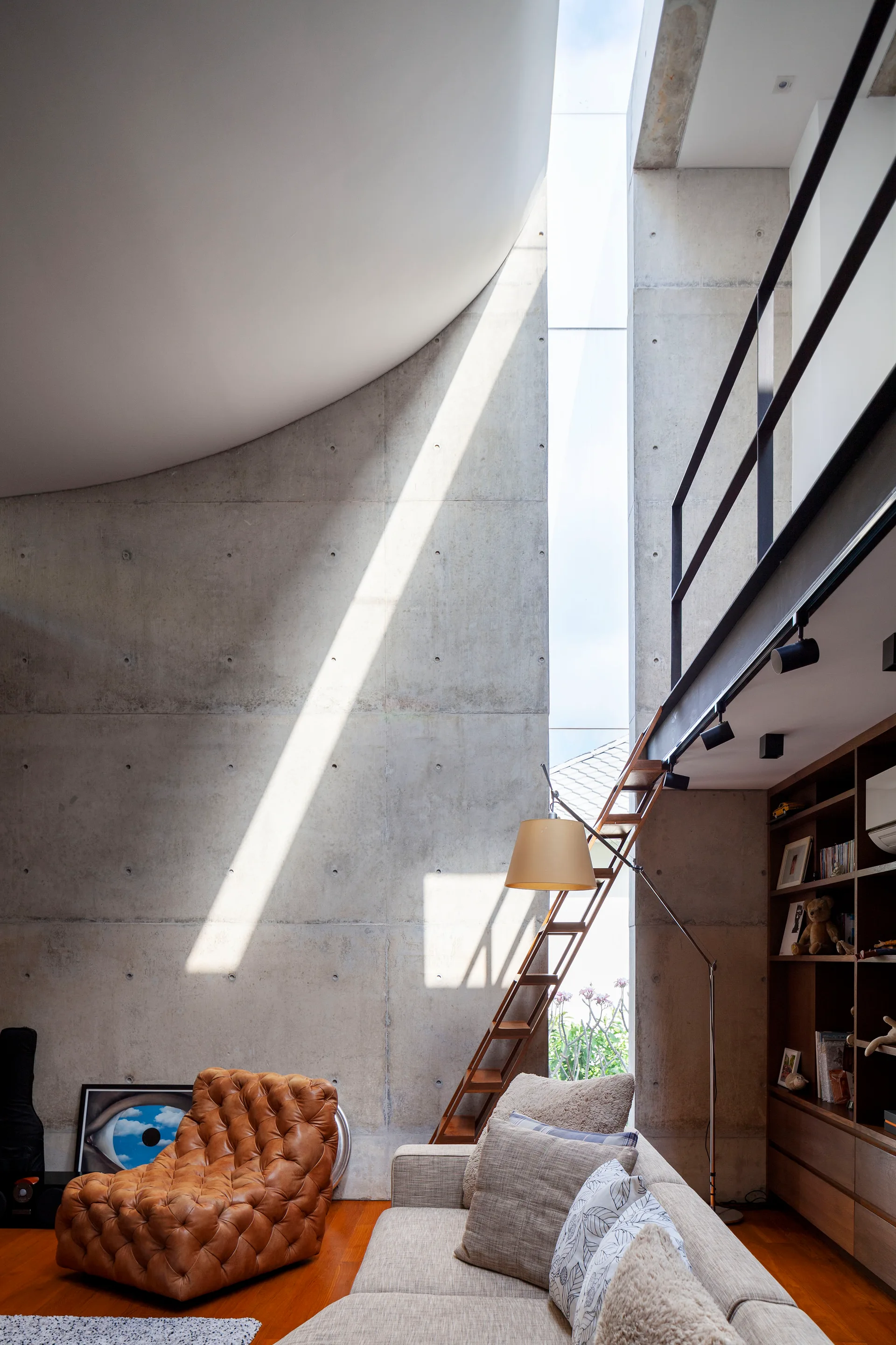 A modern living room features a tall exposed concrete wall, a vertical window letting in sunlight, a mezzanine balcony, and a tufted leather chair.