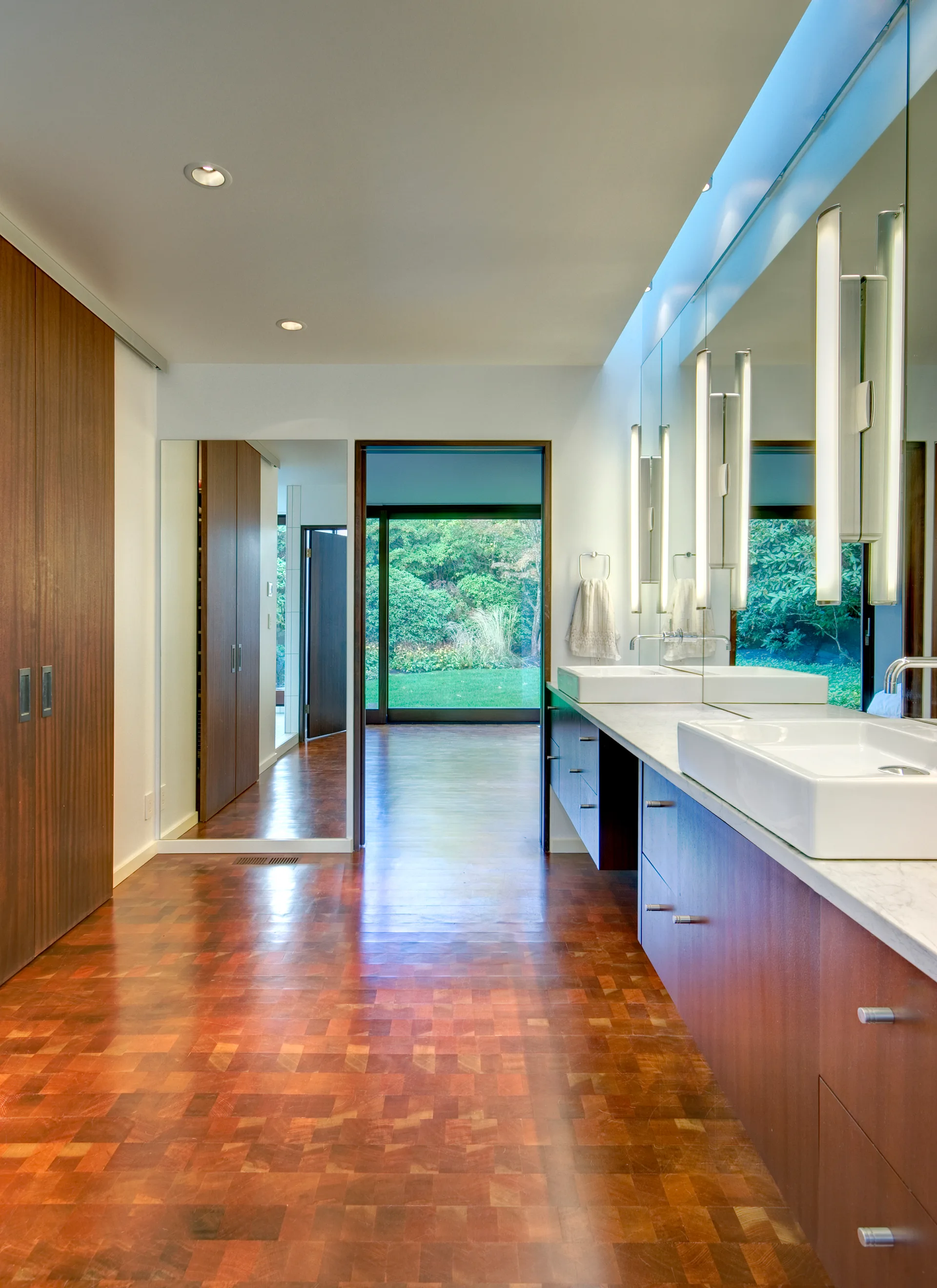A modern bathroom with a long wood vanity, white sinks, vertical wall-mounted mirrors, and parquet wood flooring leading to a view of a lush green garden.