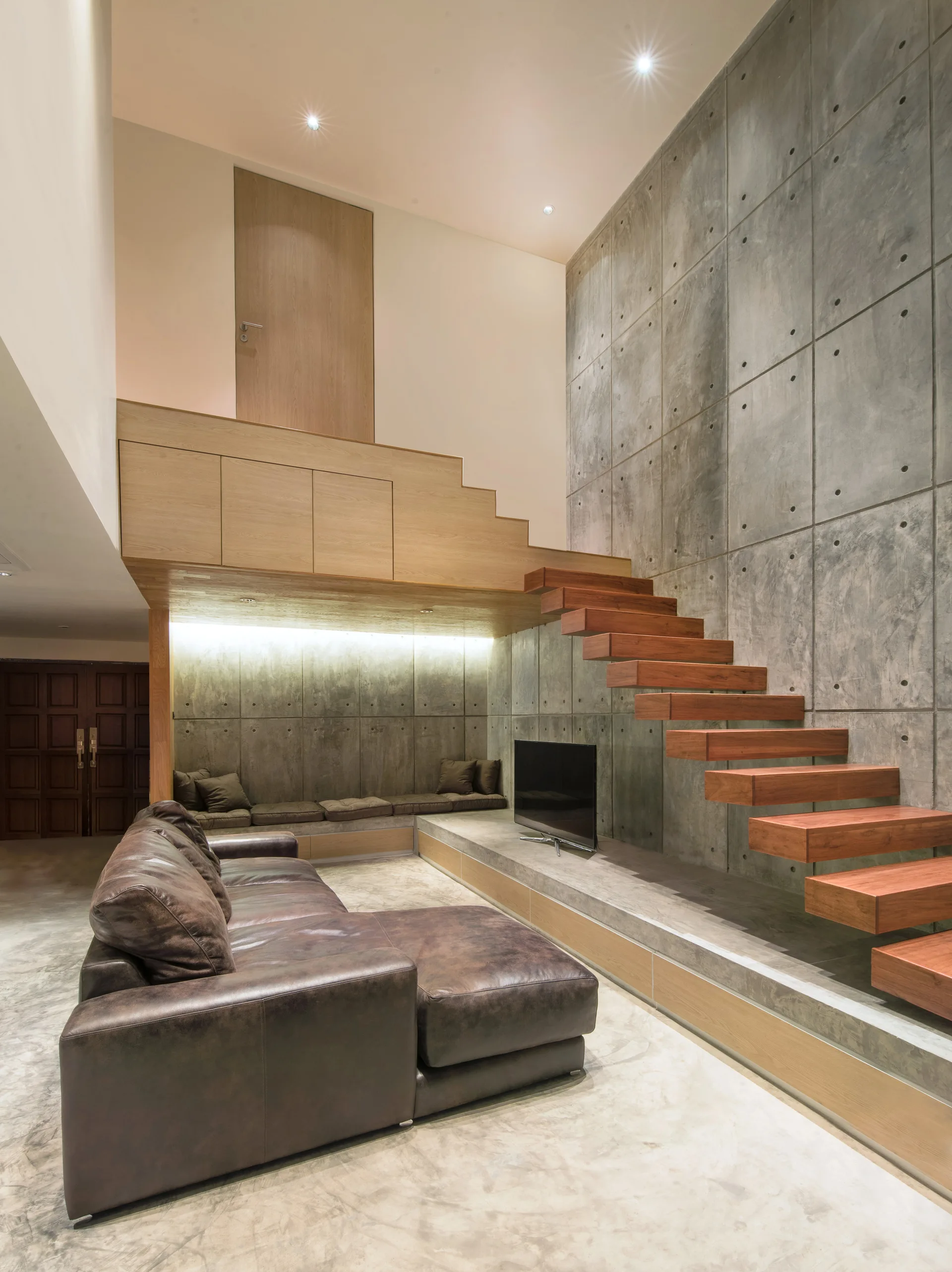 A modern living room features a brown leather sofa, a floating wooden staircase, and gridded concrete wall panels.