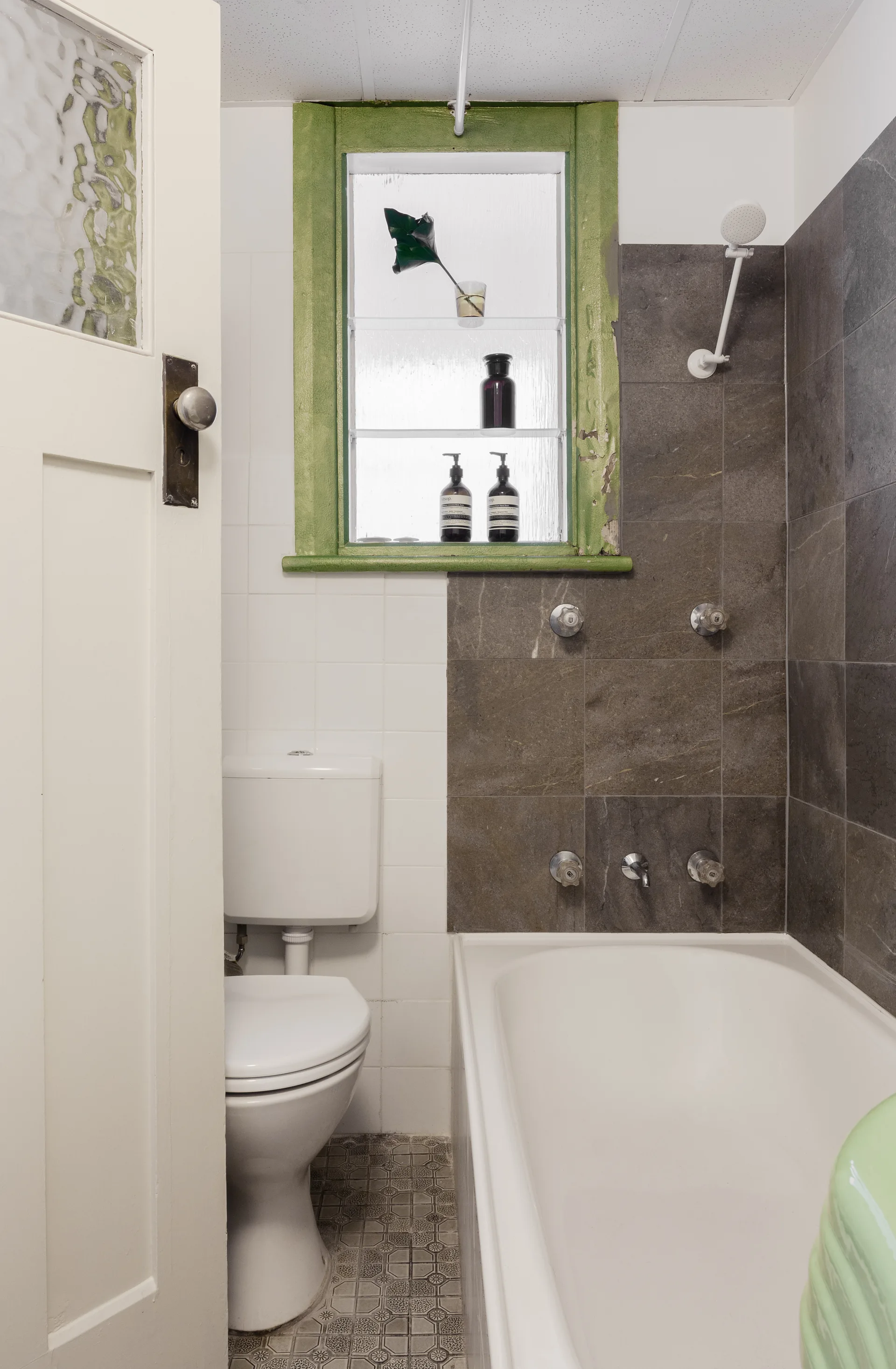 A compact bathroom features a white bathtub, a toilet, split white and dark grey wall tiles, and a green-framed window with glass shelves.