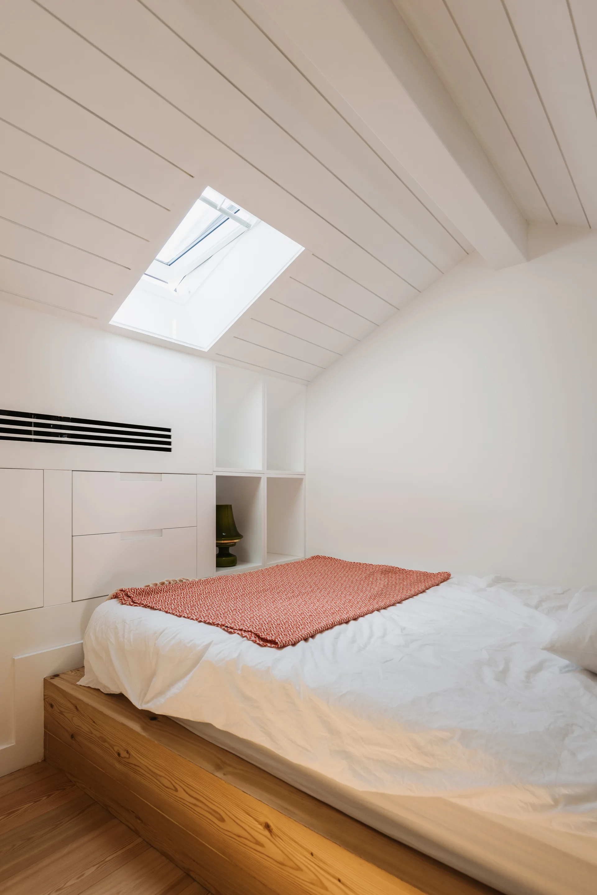 A compact attic bedroom features a low wooden platform bed under a sloped ceiling with a skylight and white built-in storage drawers.