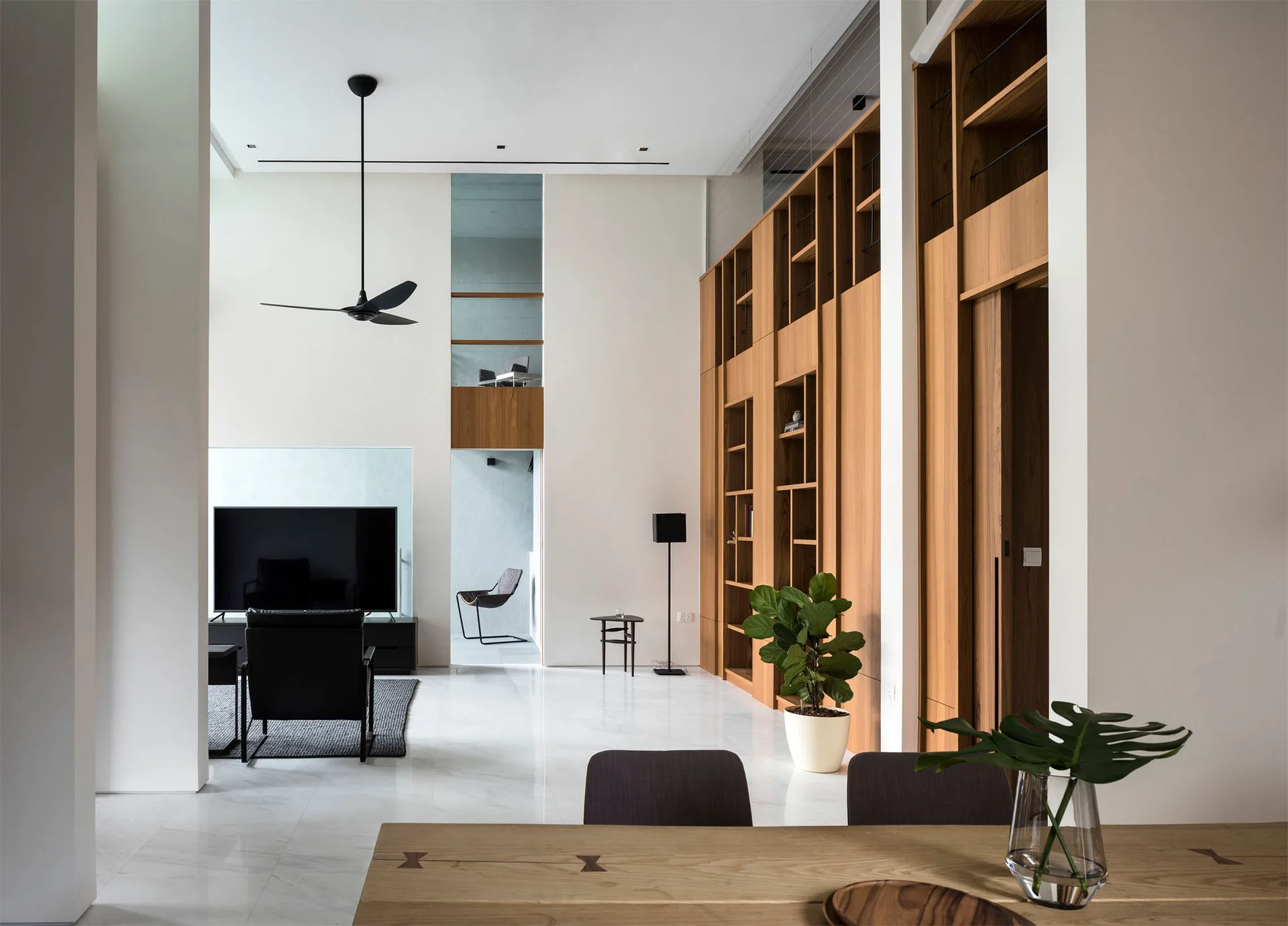 A modern living room features a tall wood storage wall, white marble flooring, black modern armchairs, and a black ceiling fan under a high ceiling.