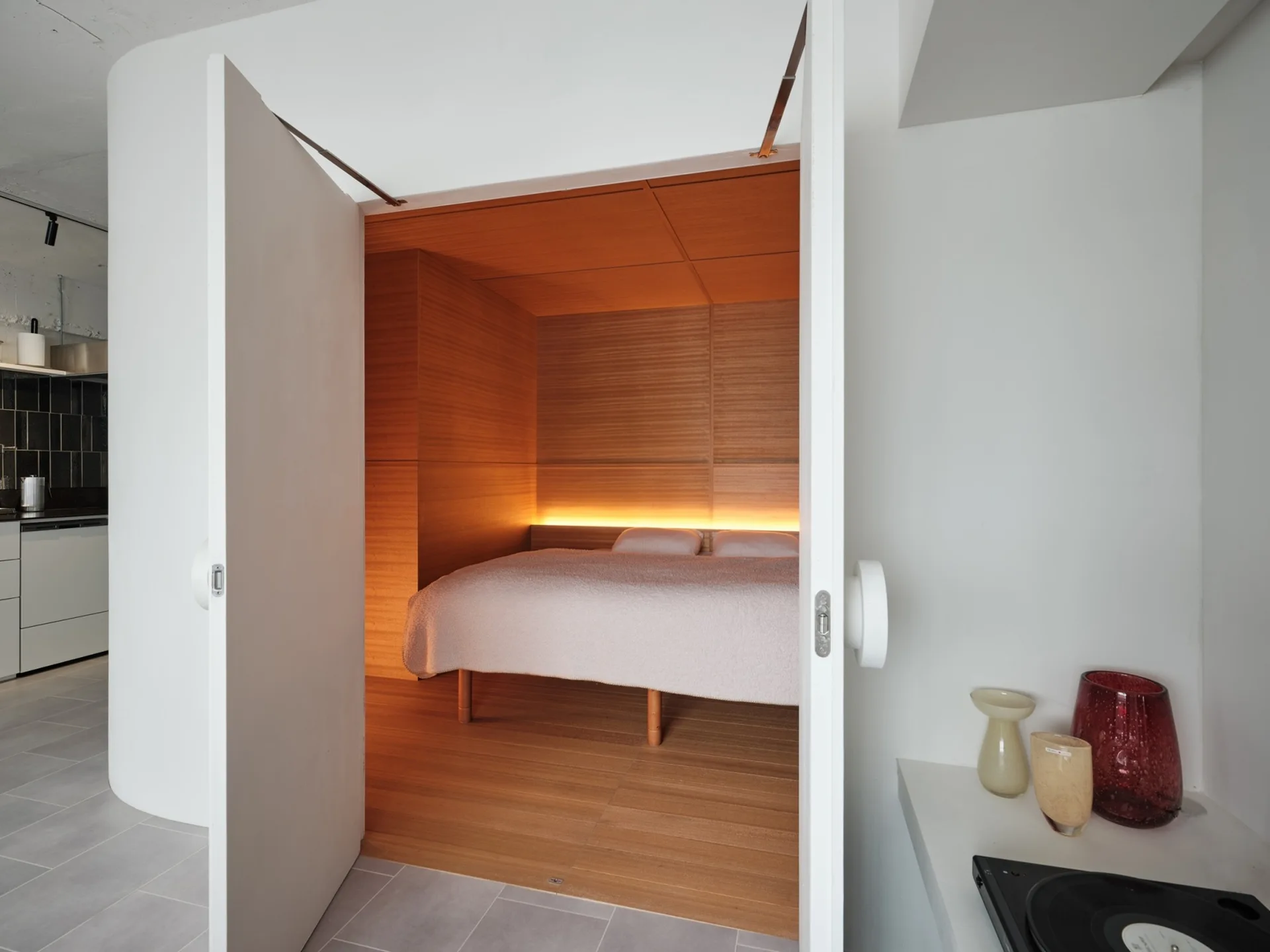 White double doors open to reveal a cozy sleeping alcove lined with warm wood paneling and illuminated by glowing headboard lighting.