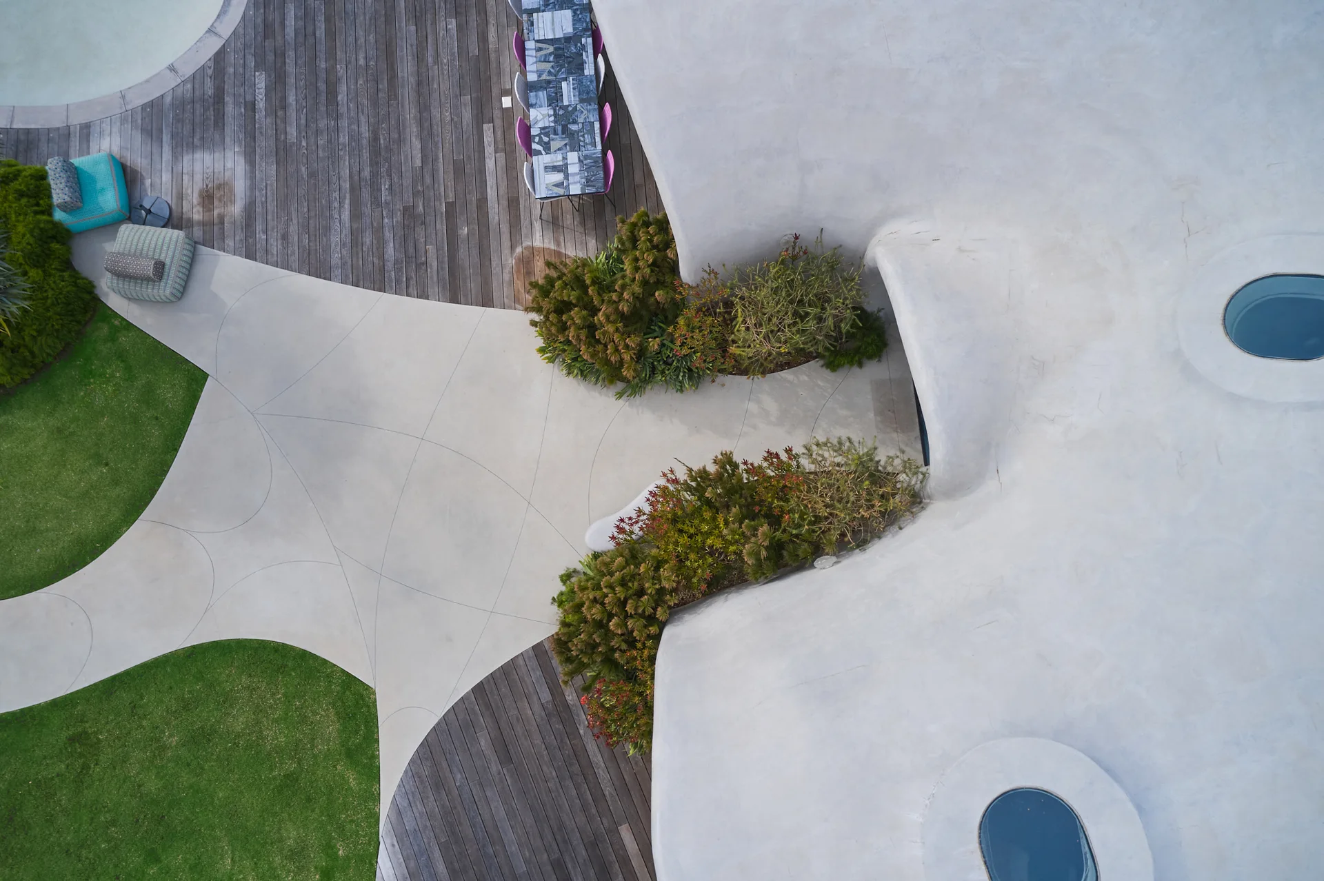 An aerial view of an organic modern patio featuring curved concrete walkways, natural wood decking, pocket gardens, and a light gray roof structure.
