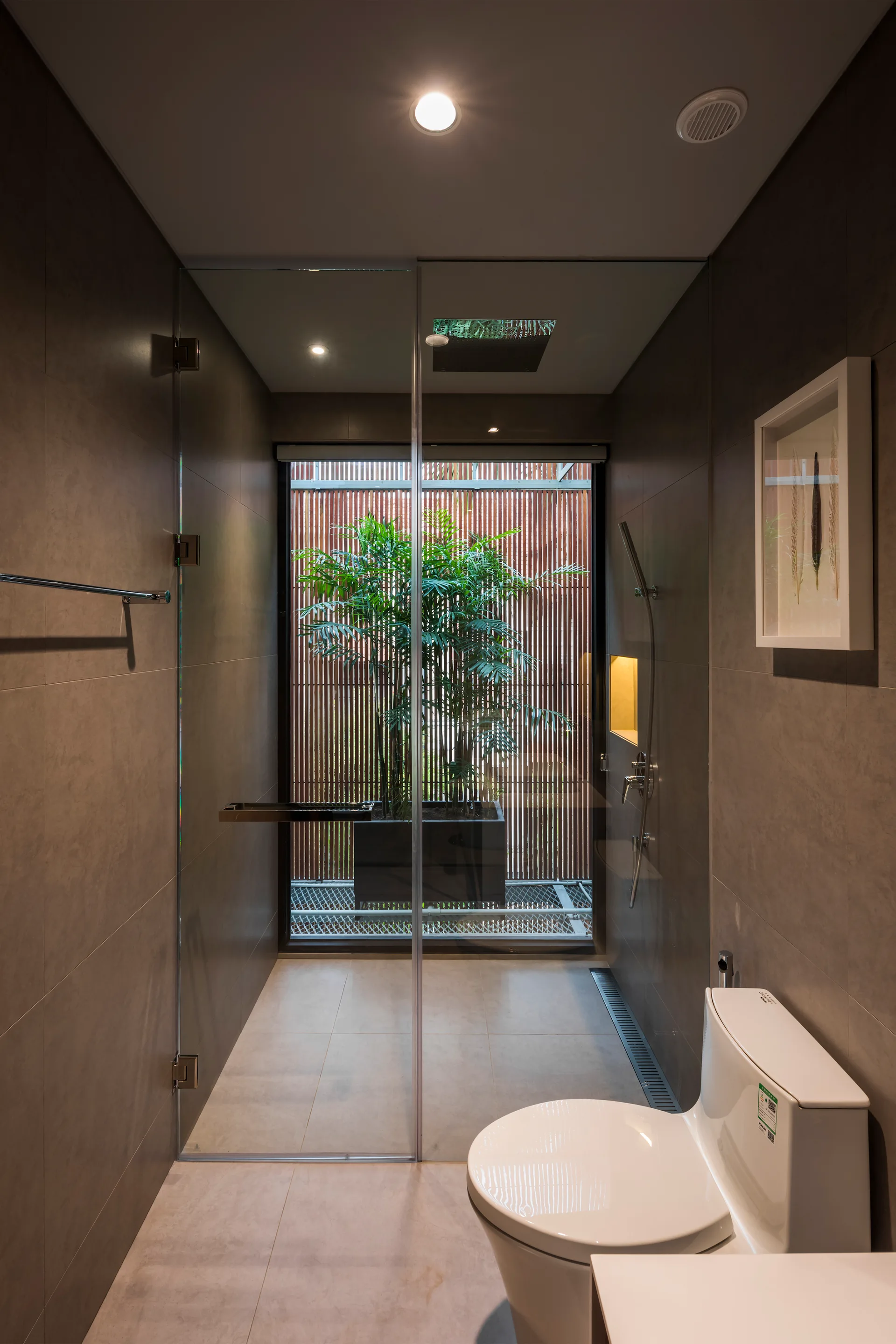 A modern gray tiled bathroom featuring a walk-in glass shower enclosure next to a toilet, looking out toward a wooden slatted privacy screen with a plant.