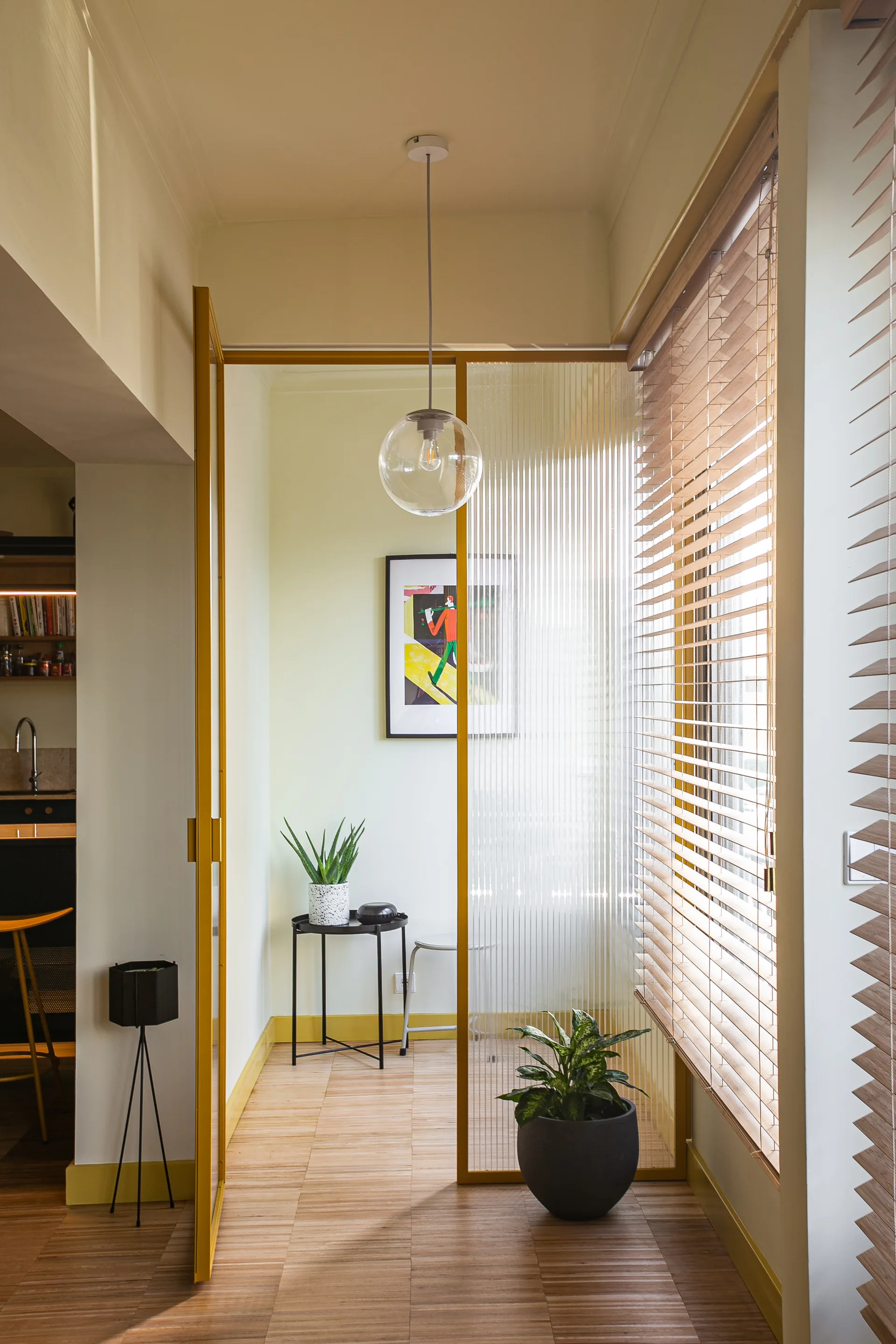 A sunlit room features a fluted glass room divider with a yellow metal frame, wooden floors, slatted wood blinds, and a globe pendant light.