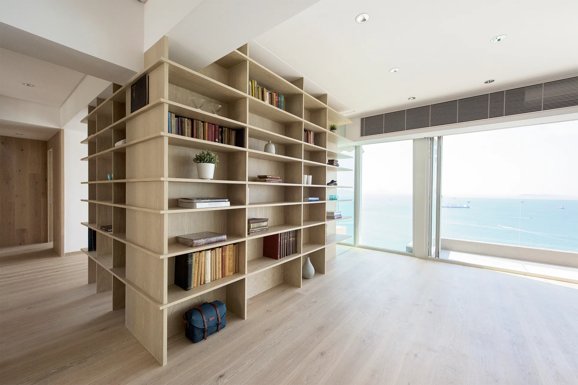 A large light wood open bookshelf wraps around a wall corner in a bright room with light wood floors and a glass window showing the ocean.