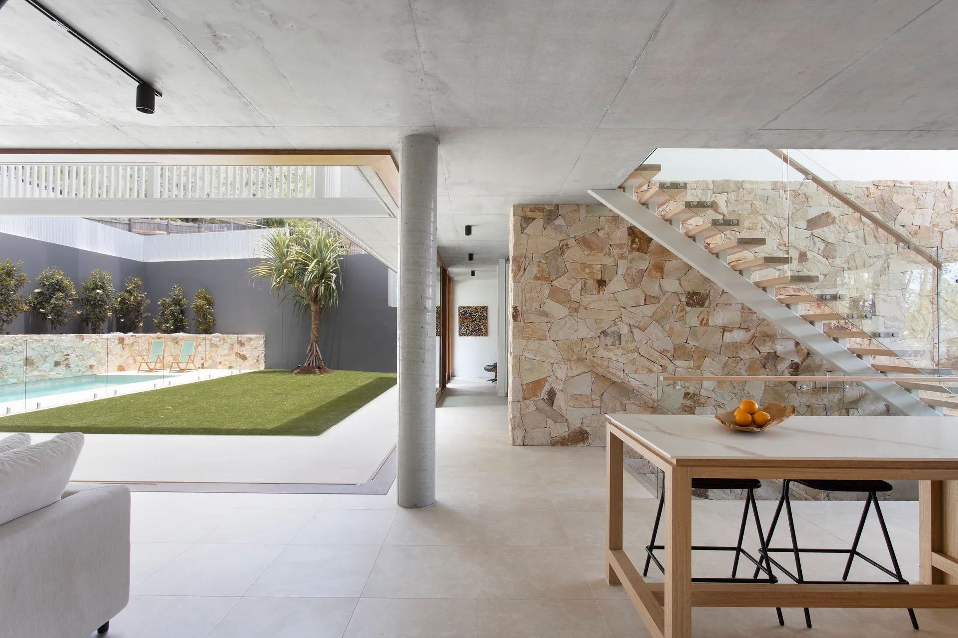 An open-plan interior features a concrete ceiling, a stone feature wall with a floating wooden staircase, and large sliding doors leading to a pool patio.