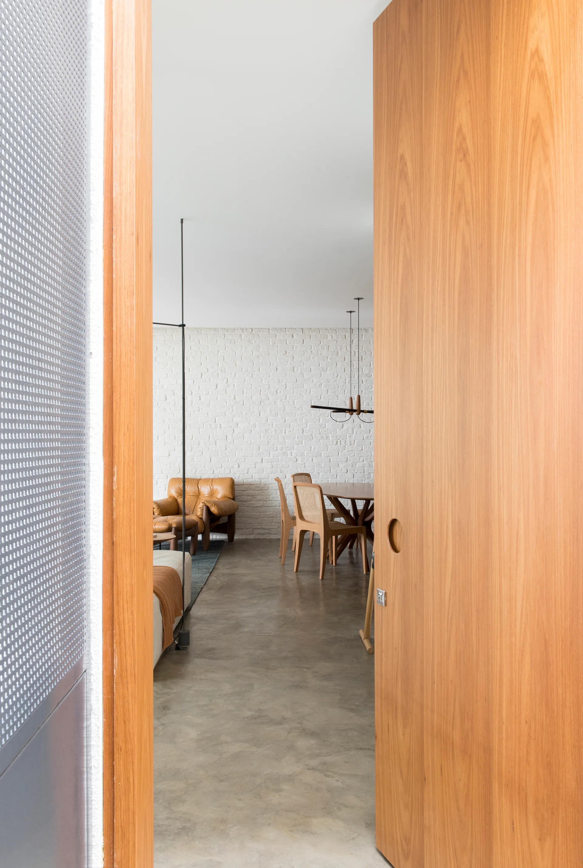 A large wooden pivot door opens to reveal a living and dining room with a white-painted brick wall, polished concrete floor, and mid-century furniture.