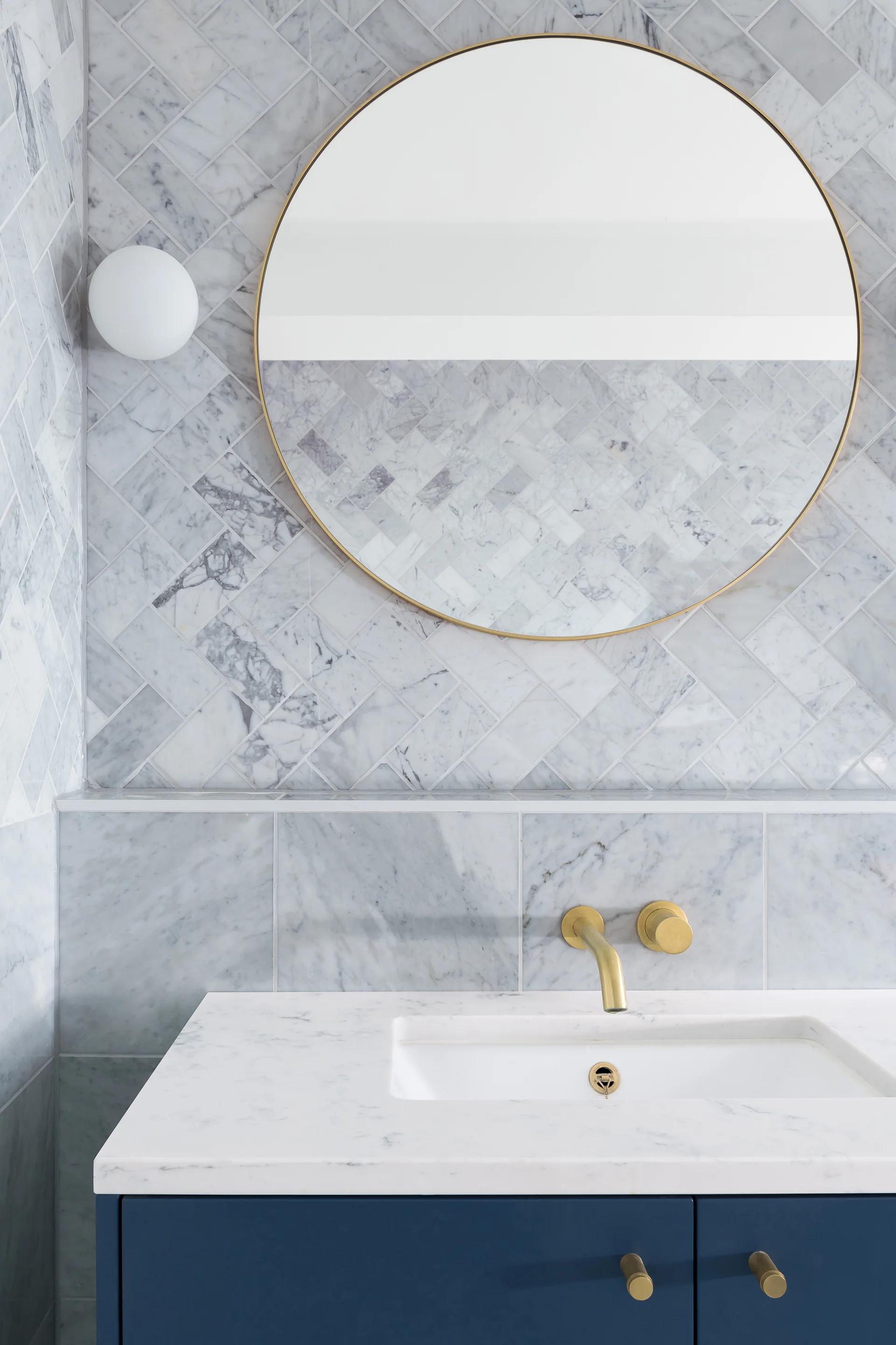 A navy bathroom vanity with a marble countertop, round brass mirror, and wall-mounted brass faucet against herringbone marble tile.
