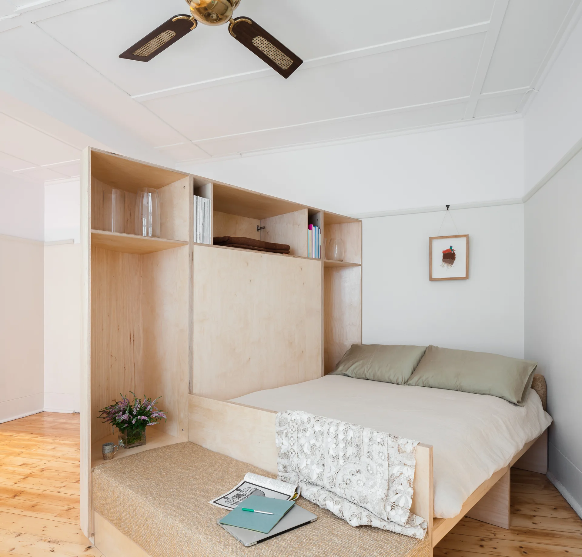 A bed with sage green pillows is nestled against a custom light wood plywood room divider featuring open shelves and an integrated bench.
