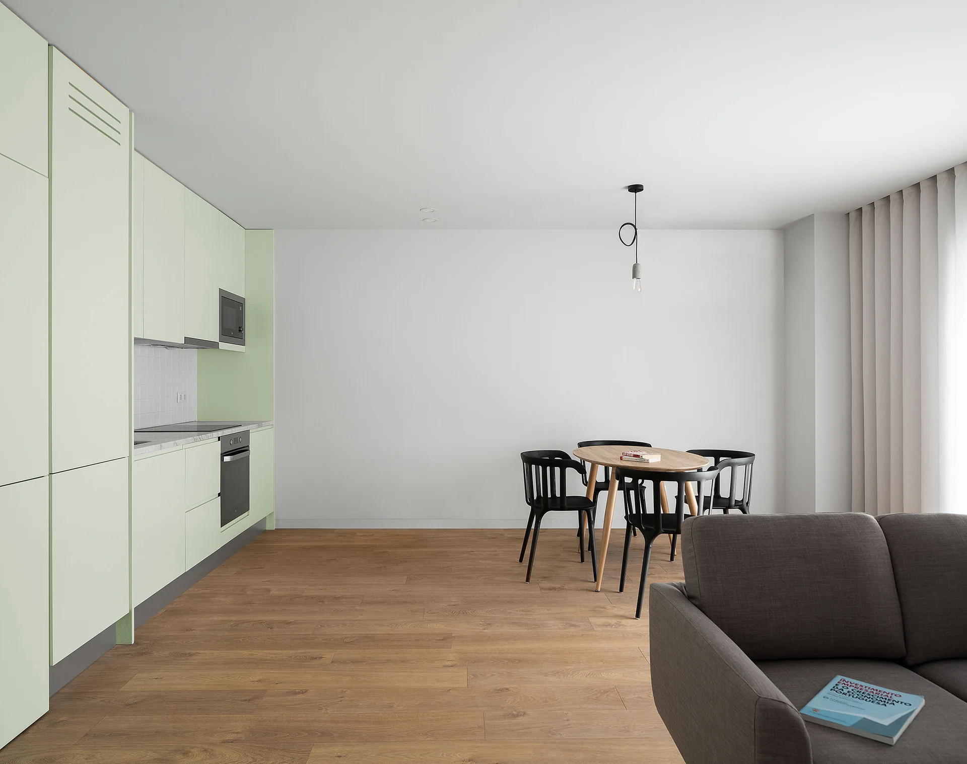 A modern open-plan interior features flat-front pale green kitchen cabinets, a round wooden dining table with four black chairs, and a grey sofa.