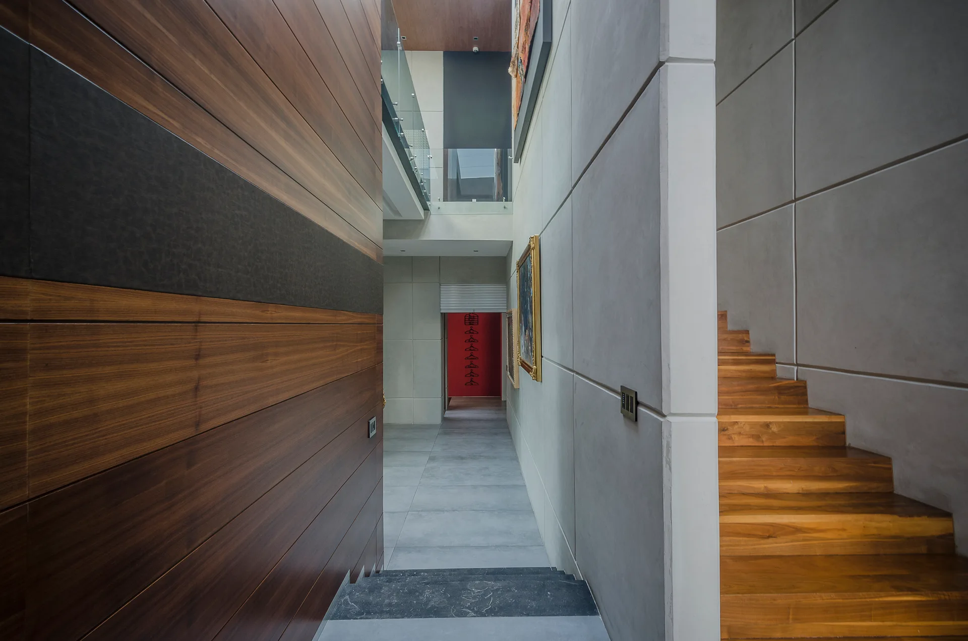 A modern hallway features concrete wall panels, a wooden staircase on the right, and a glimpse of a red accent wall in the distance.