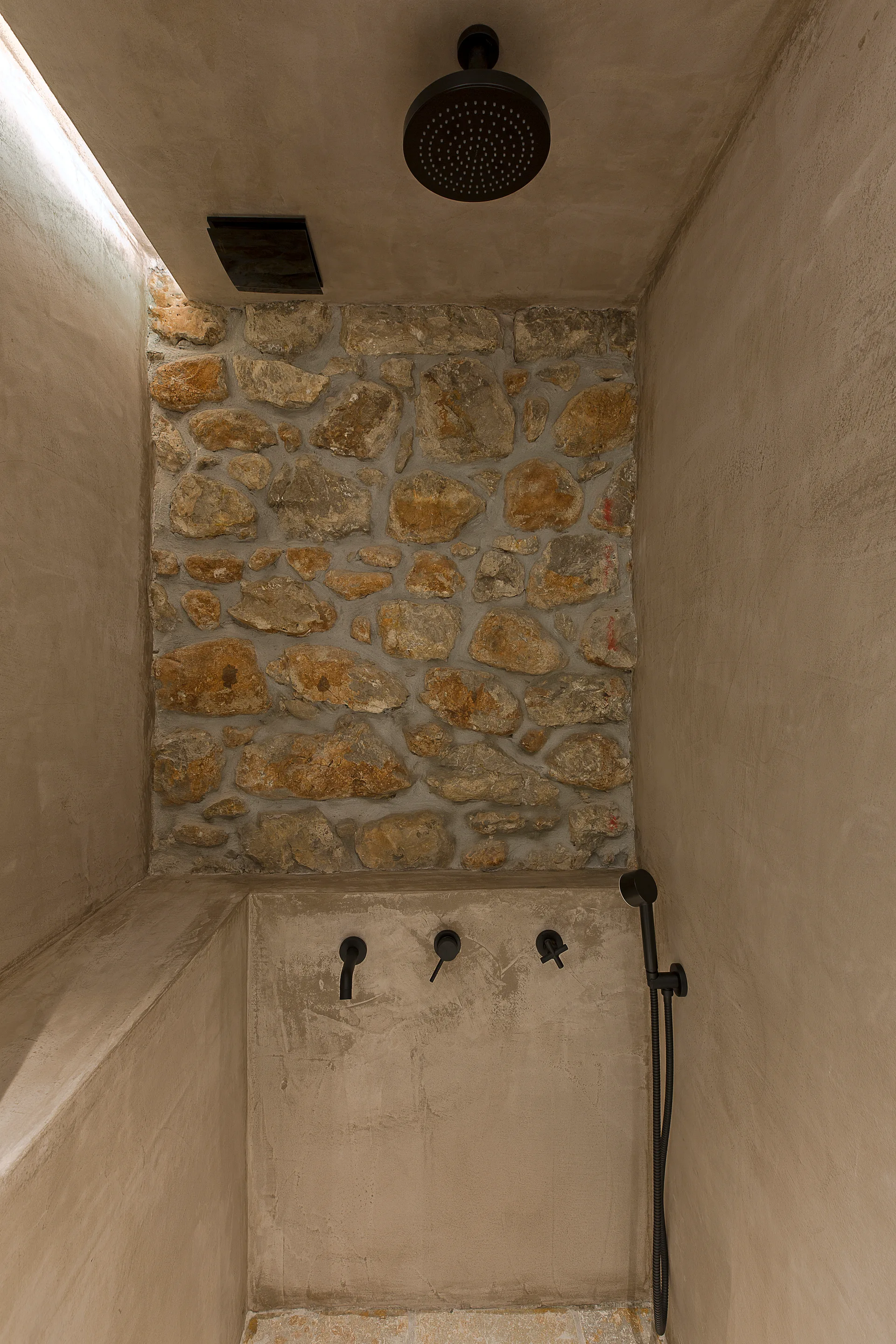 An organic modern shower features a textured natural stone accent wall surrounded by smooth beige plaster and minimalist matte black hardware.