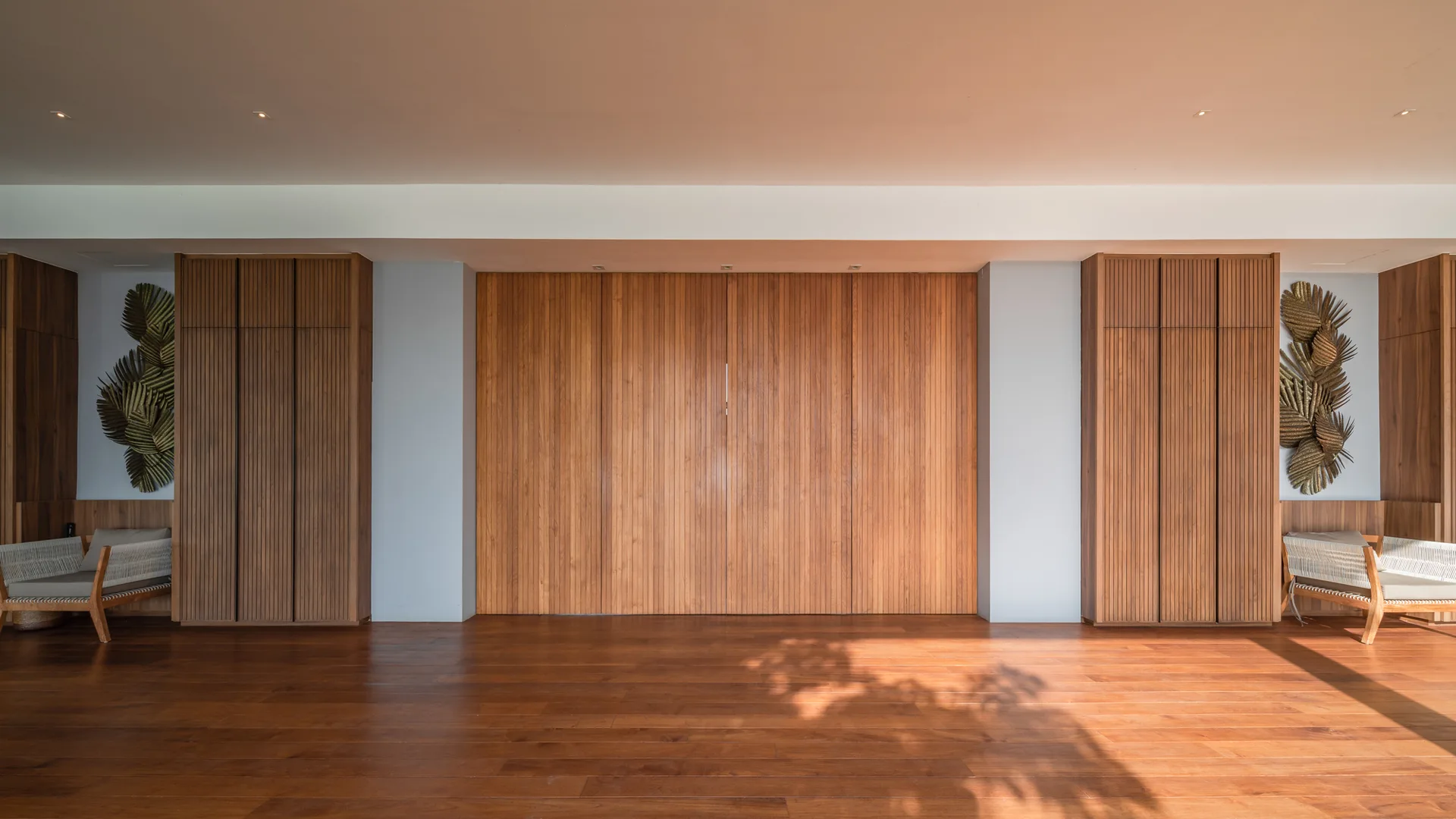 A symmetrical room features a floor-to-ceiling wood paneled wall, integrated slatted wood cabinets, and two low-profile armchairs on hardwood floors.