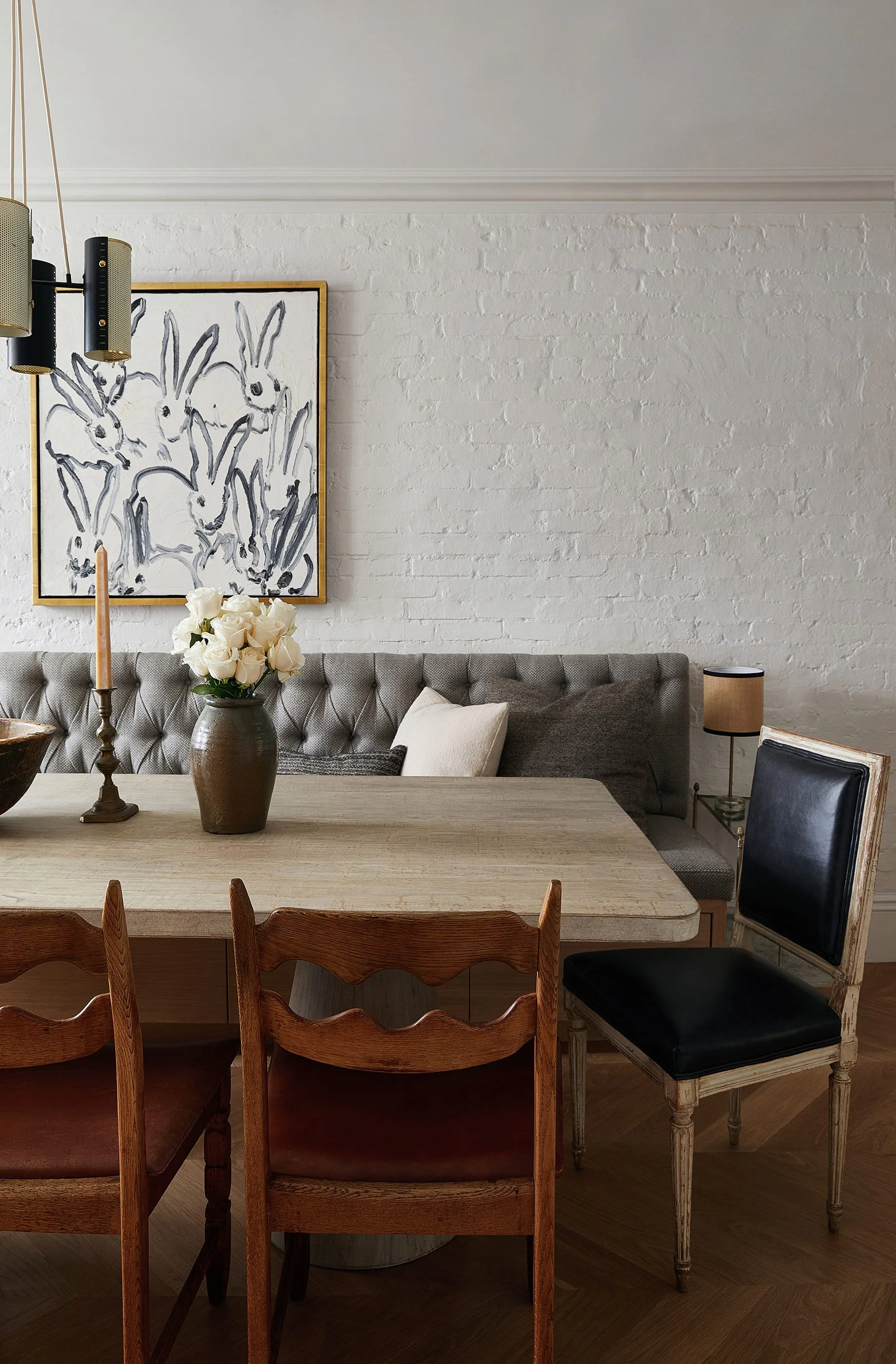 A dining room features a light wood table, a grey tufted banquette against a white brick wall, and mismatched wooden chairs on herringbone flooring.